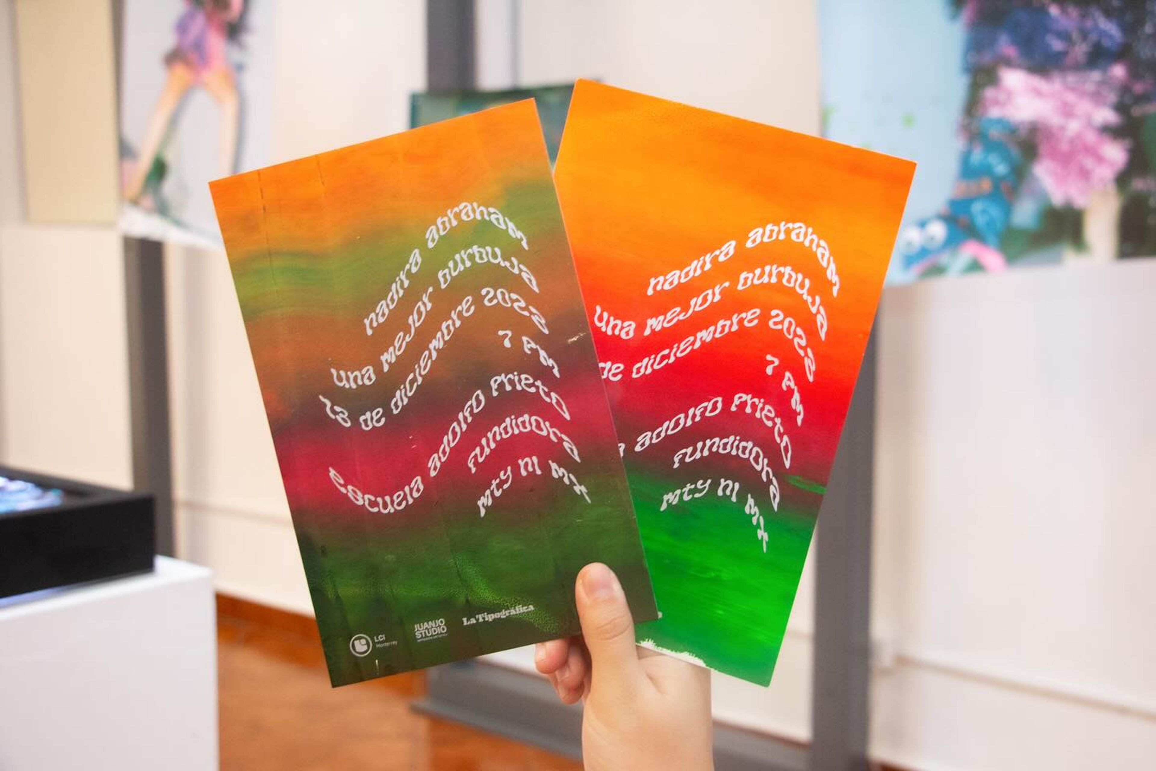 Manos sosteniendo dos folletos de evento vibrantes con gradientes de color y texto, promocionando una exposición de arte, ejemplificando el marketing creativo en las artes.