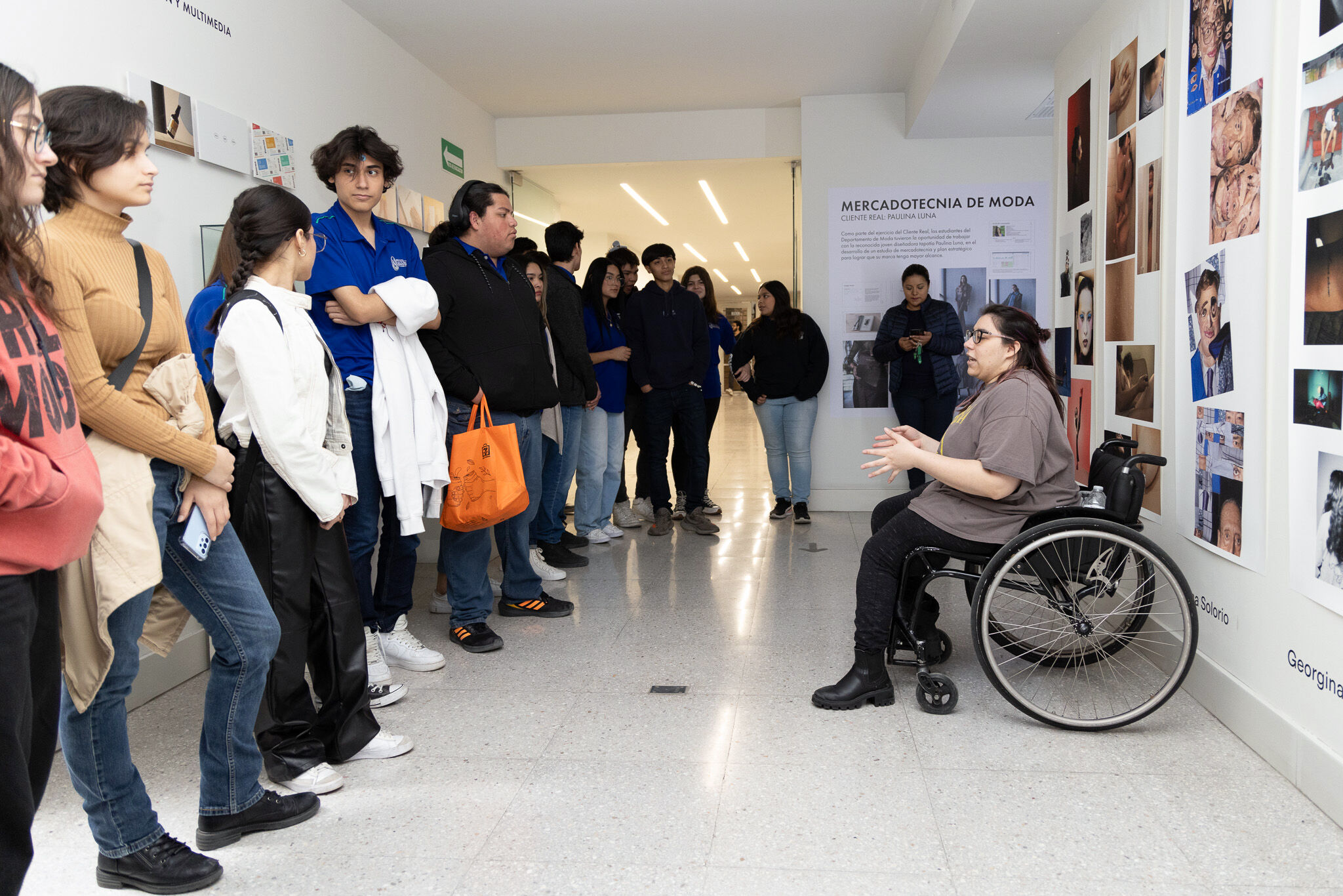Un grupo diverso de estudiantes escucha atentamente a un presentador en silla de ruedas en un evento de galería académica, rodeado de proyectos creativos en las paredes.