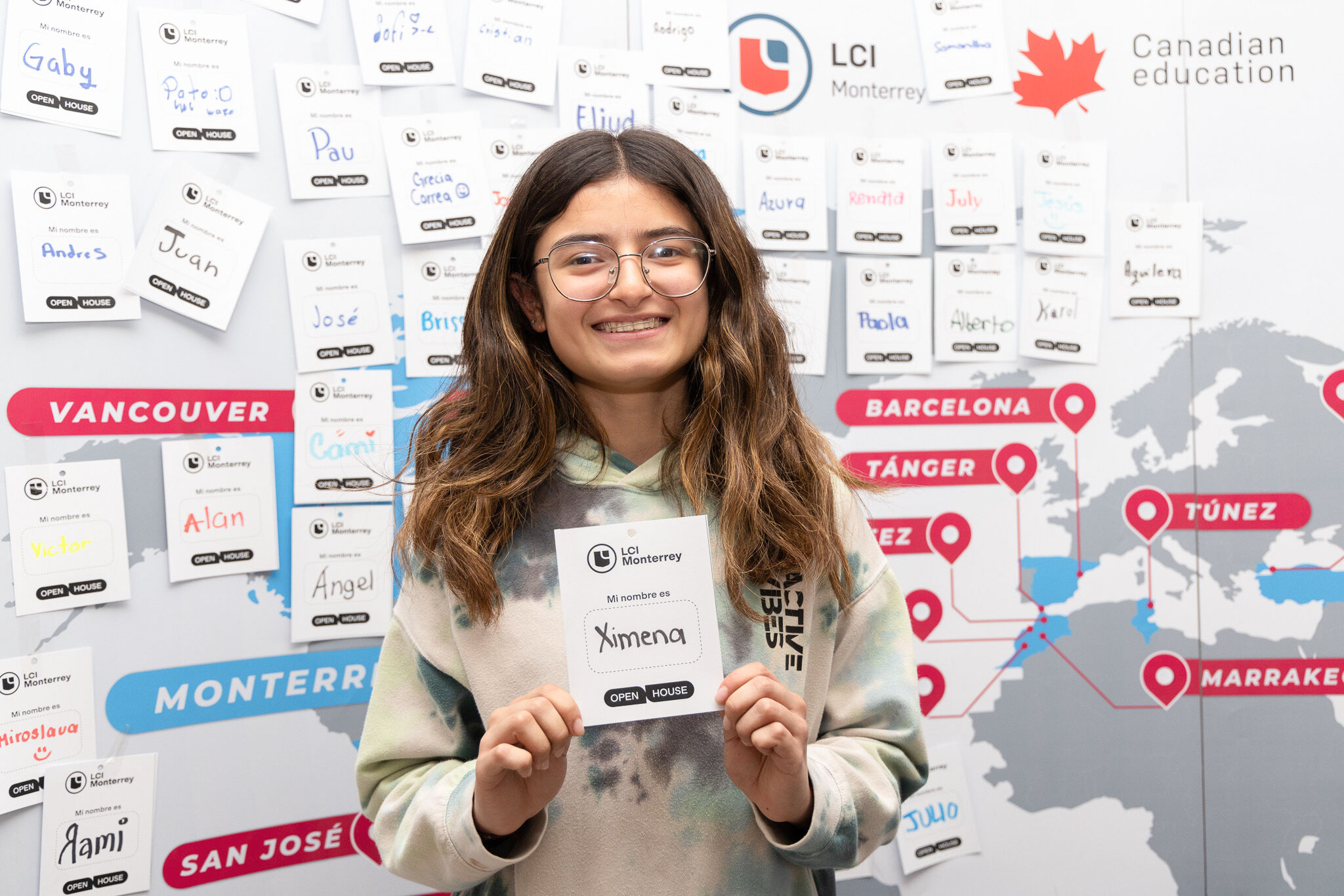 Una joven alegre sostiene su etiqueta de nombre en una jornada de puertas abiertas de una escuela internacional, con un mapa mundial y nombres de ciudades en el fondo que indican varios lugares de estudio.