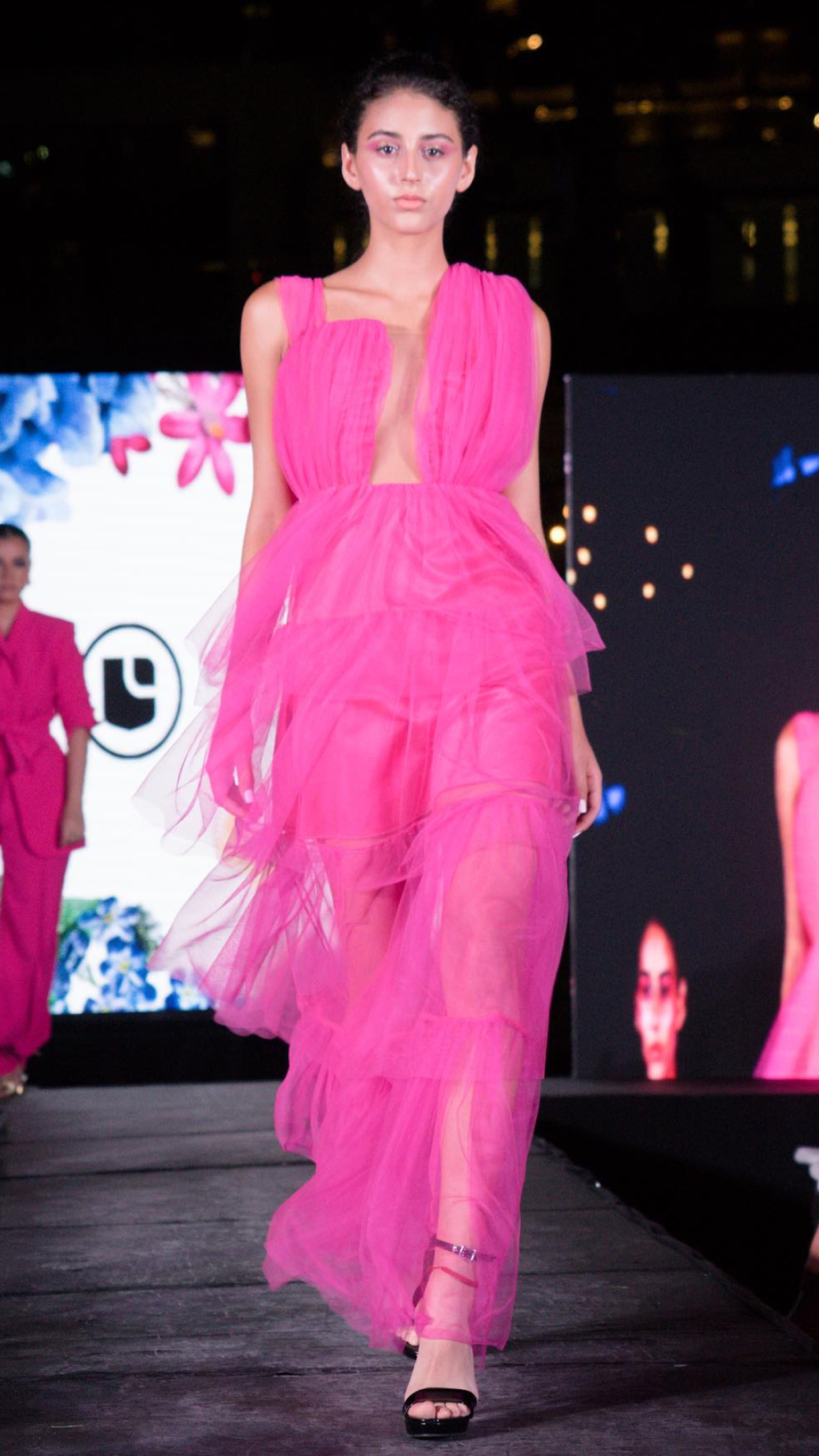 A model strides on a fashion show runway, wearing a bold pink tulle gown with a plunging neckline.
