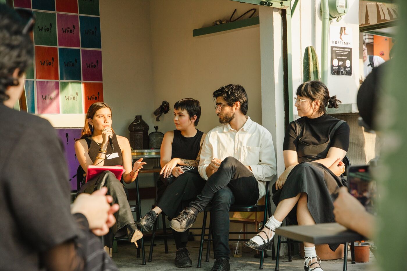 Four panelists engage in a discussion before an audience at a creative workshop, with colorful "Who?" question posters in the background.