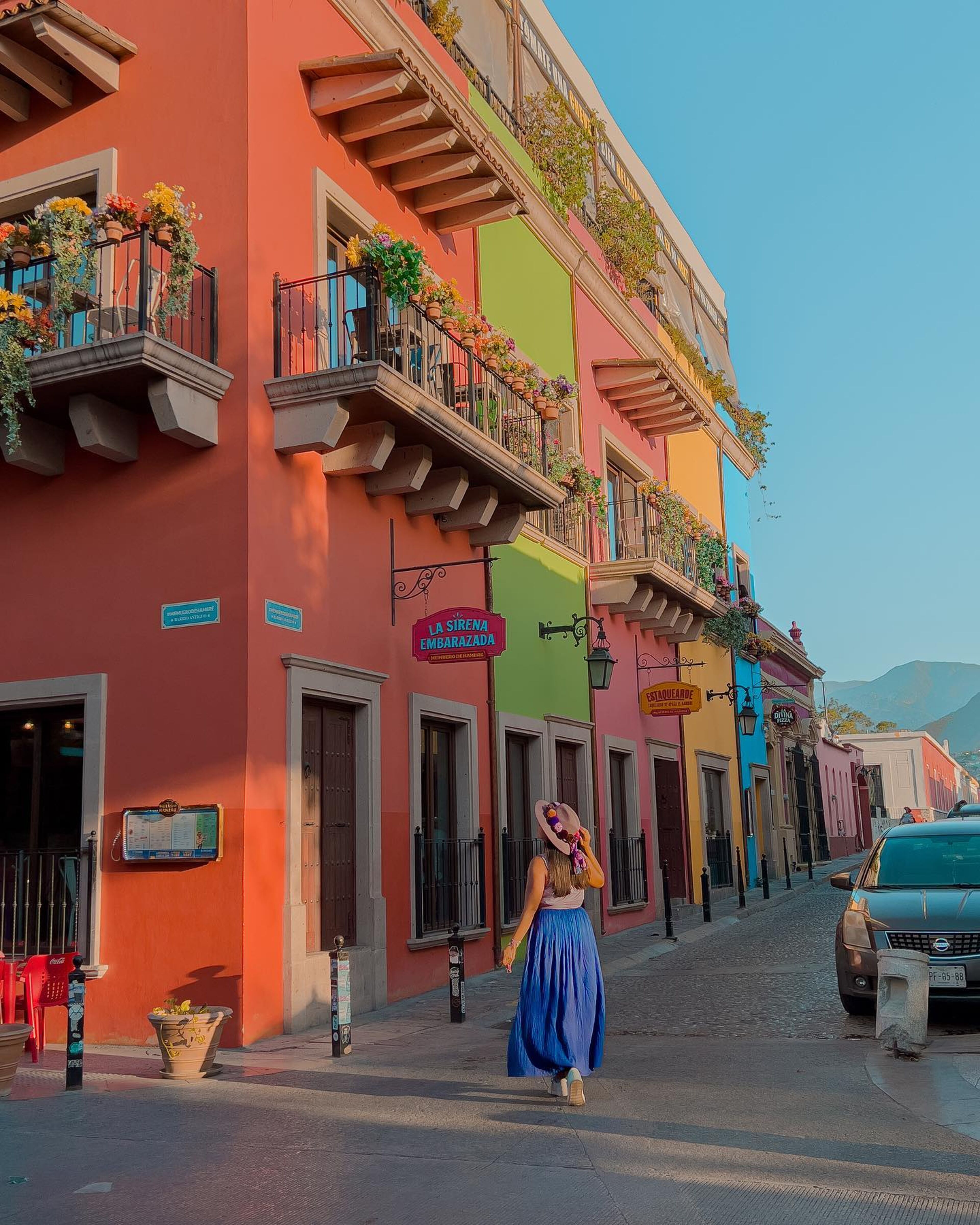 A woman in a blue skirt and hat walks down a vibrant street lined with colonial buildings in the warm glow of the late afternoon sun.

