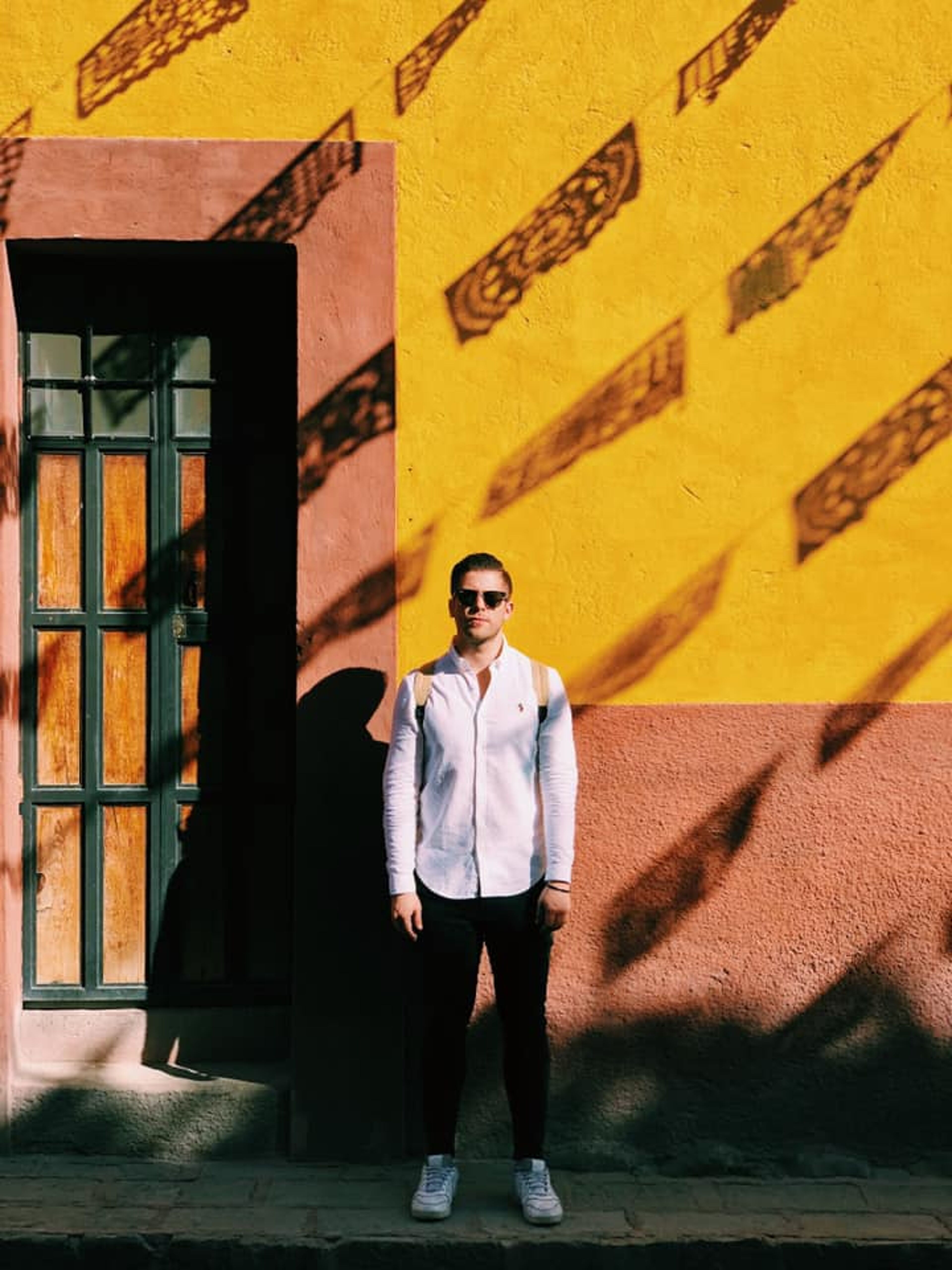 Un hombre con ropa casual está de pie frente a una pared amarilla vibrante con patrones de sombras, al lado de una puerta de madera.