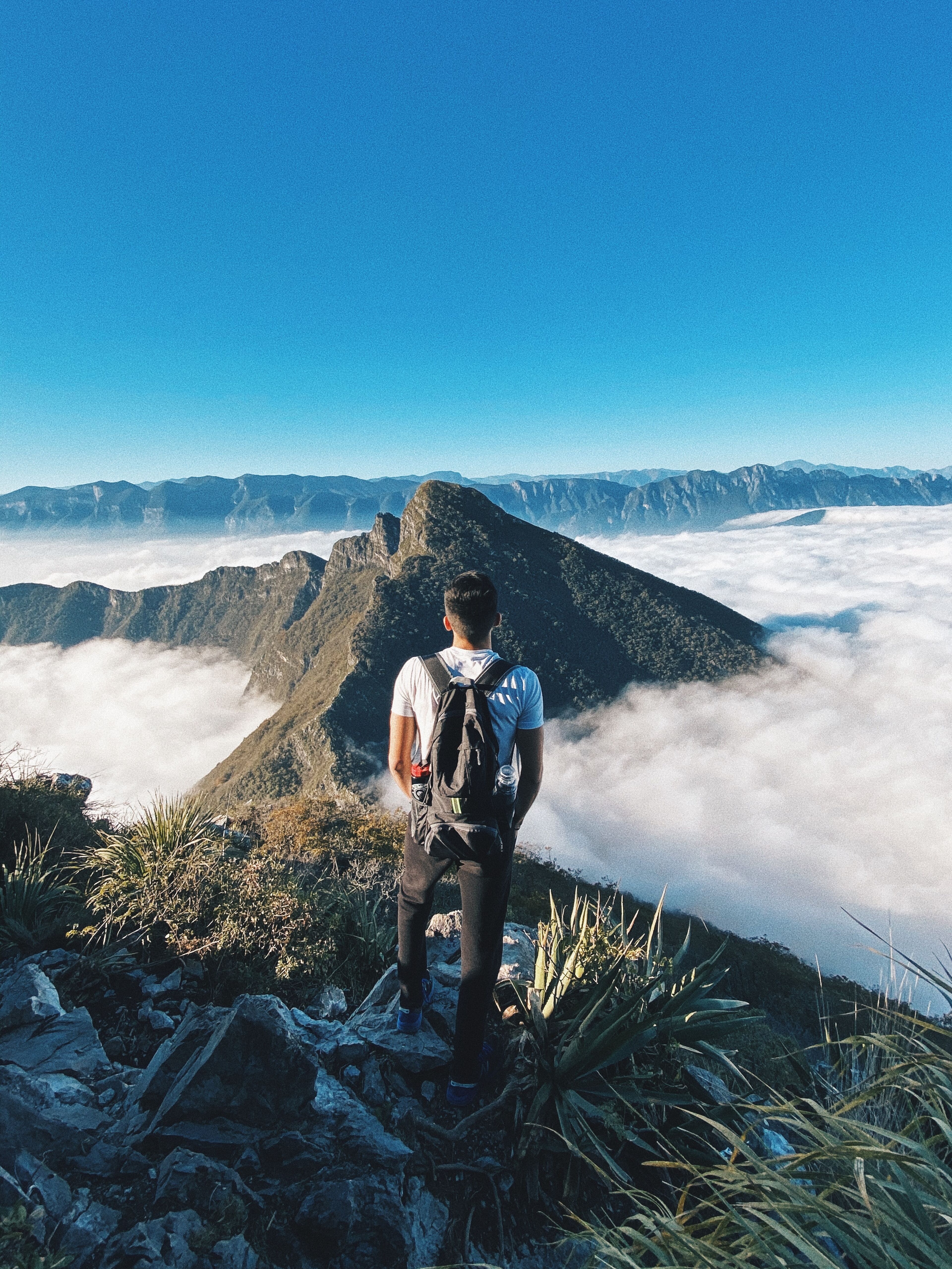 Un excursionista solitario se para en la cima de un pico accidentado, observando un mar de nubes que envuelven montañas distantes bajo un cielo azul despejado.