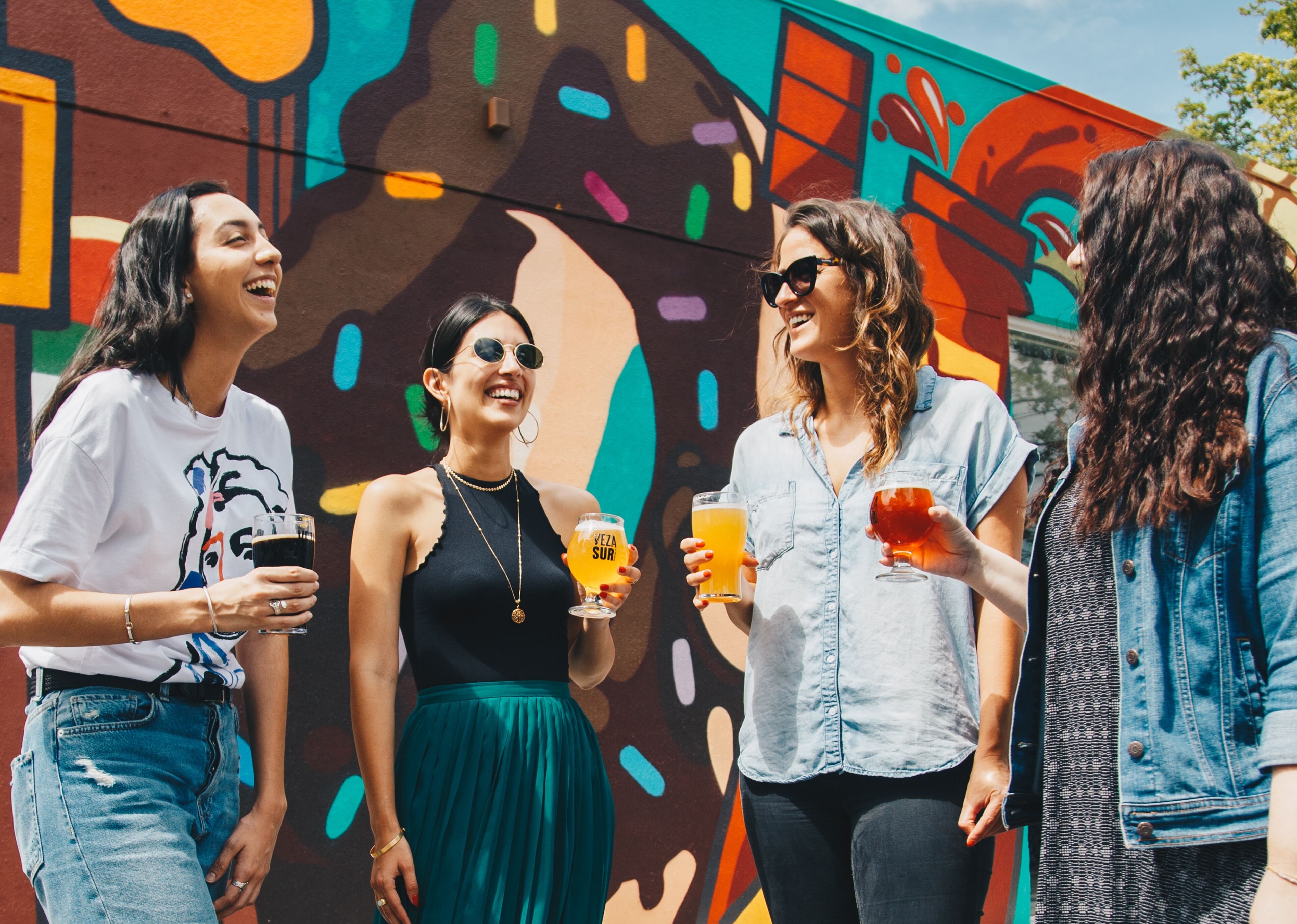 Cuatro mujeres se ríen y disfrutan de bebidas frente a un mural colorido.