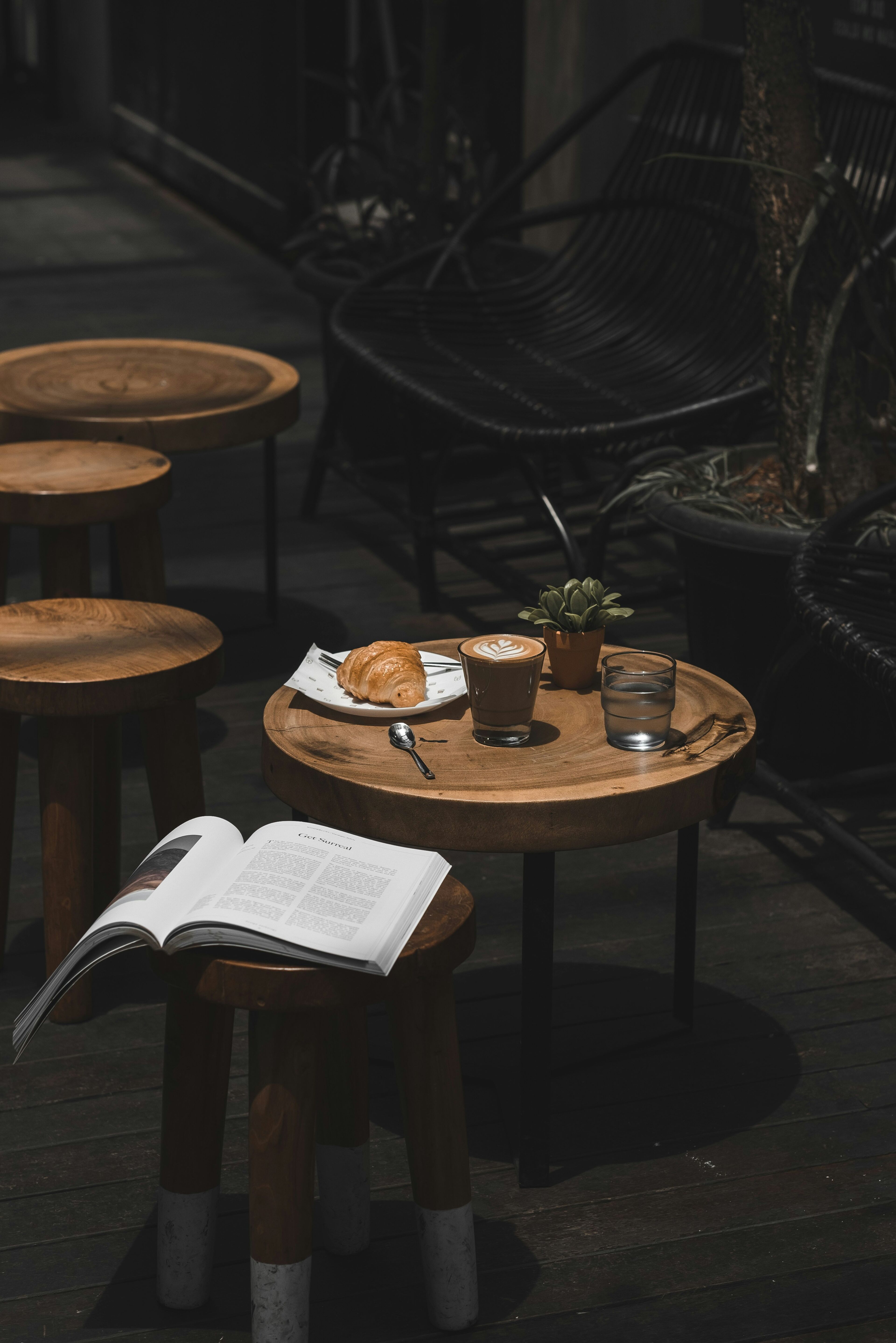 Un entorno tranquilo con una mesa de madera con una taza de café, un cruasán, un cactus y material de lectura, junto a una silla de mimbre en un patio oscuro.