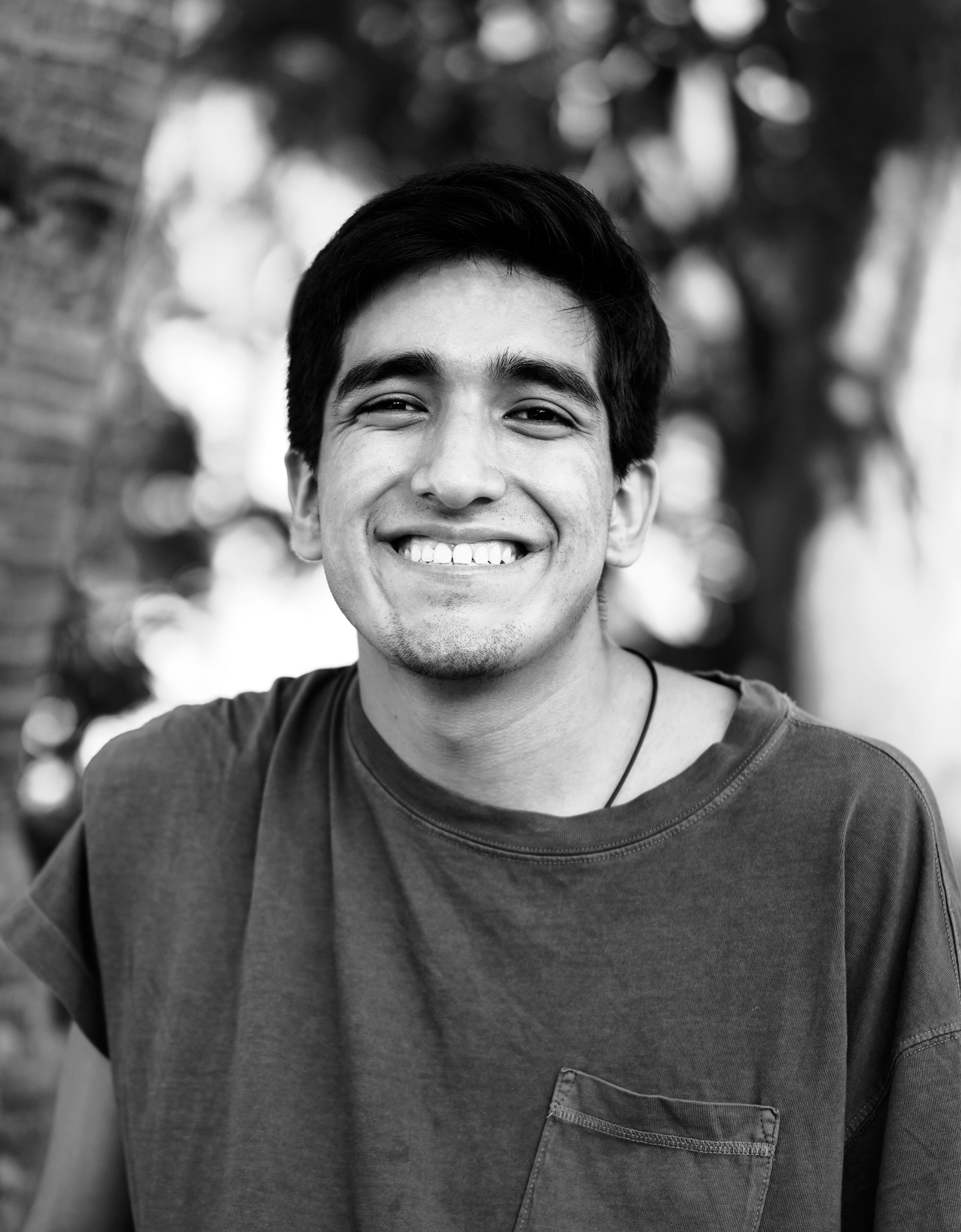 Un joven adulto masculino sonriendo ampliamente con una camiseta informal, con un fondo natural borroso que sugiere un entorno al aire libre.