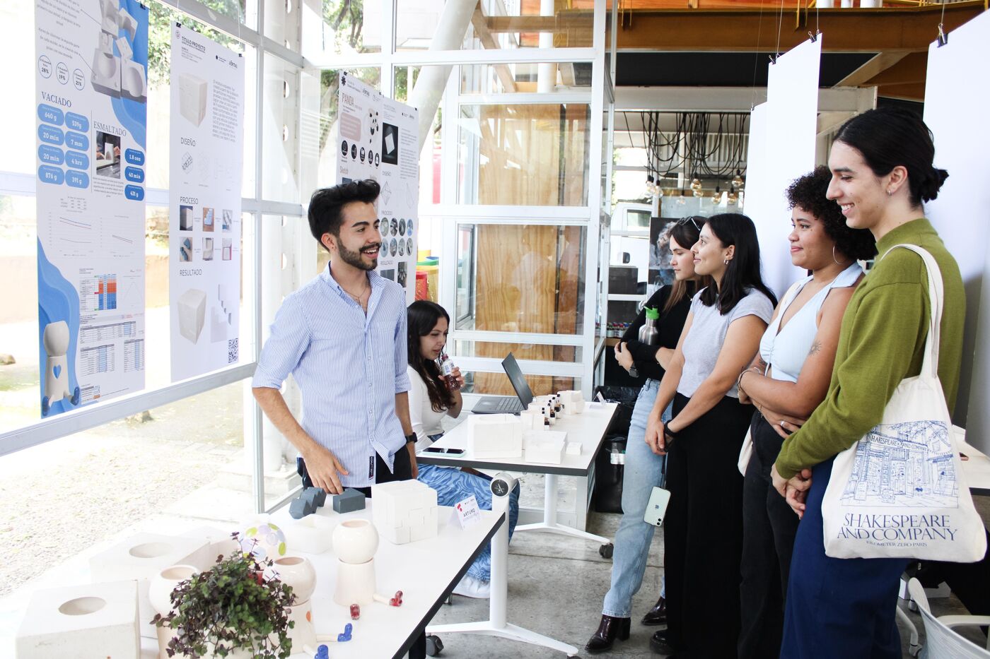 A group of young adults stand in a brightly lit room, some looking at architectural models on display tables, while others converse near project boards.