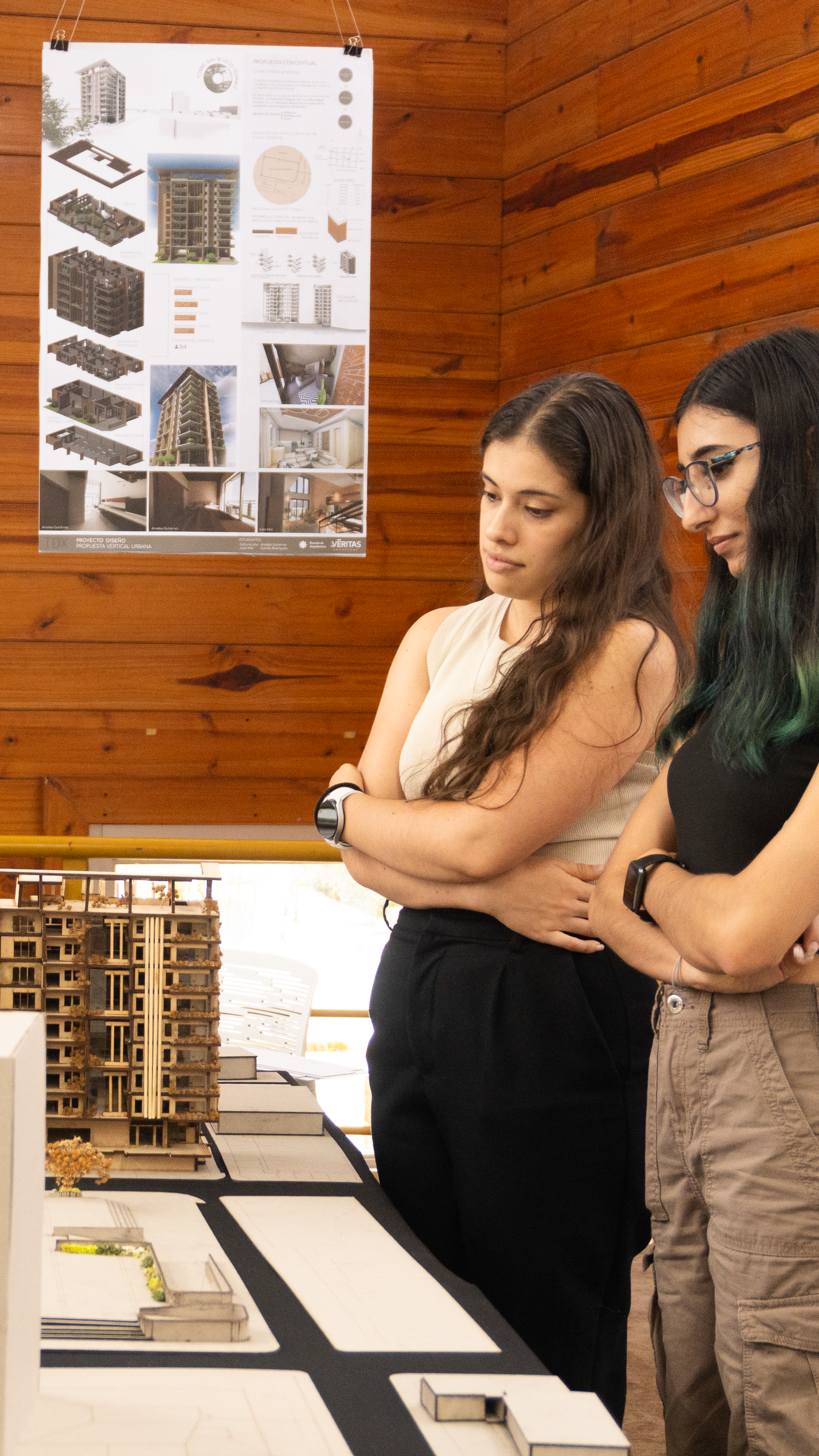 Two students closely examine architectural models and plans displayed on a table.