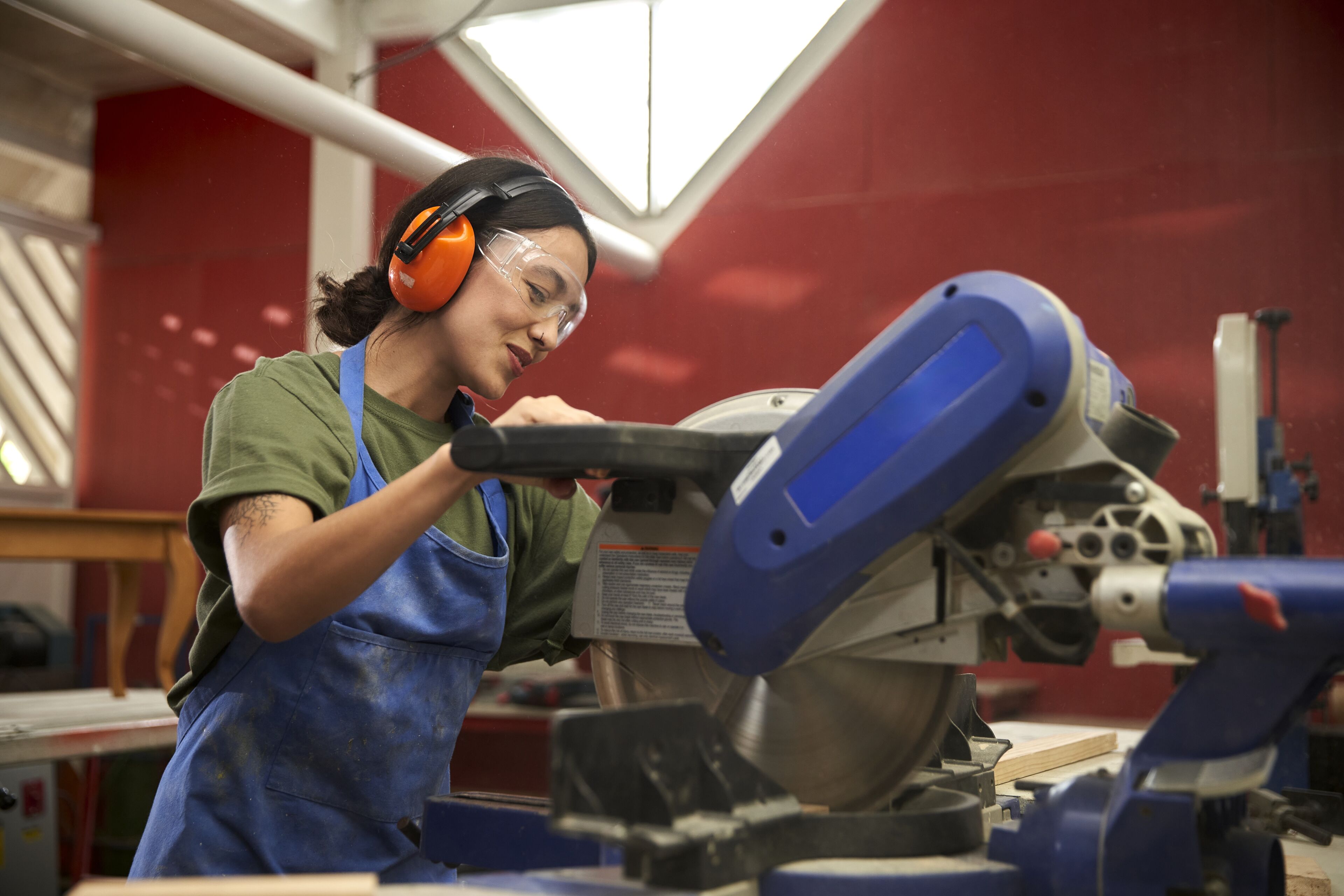 A focused craftswoman in safety gear uses a miter saw in a workshop.