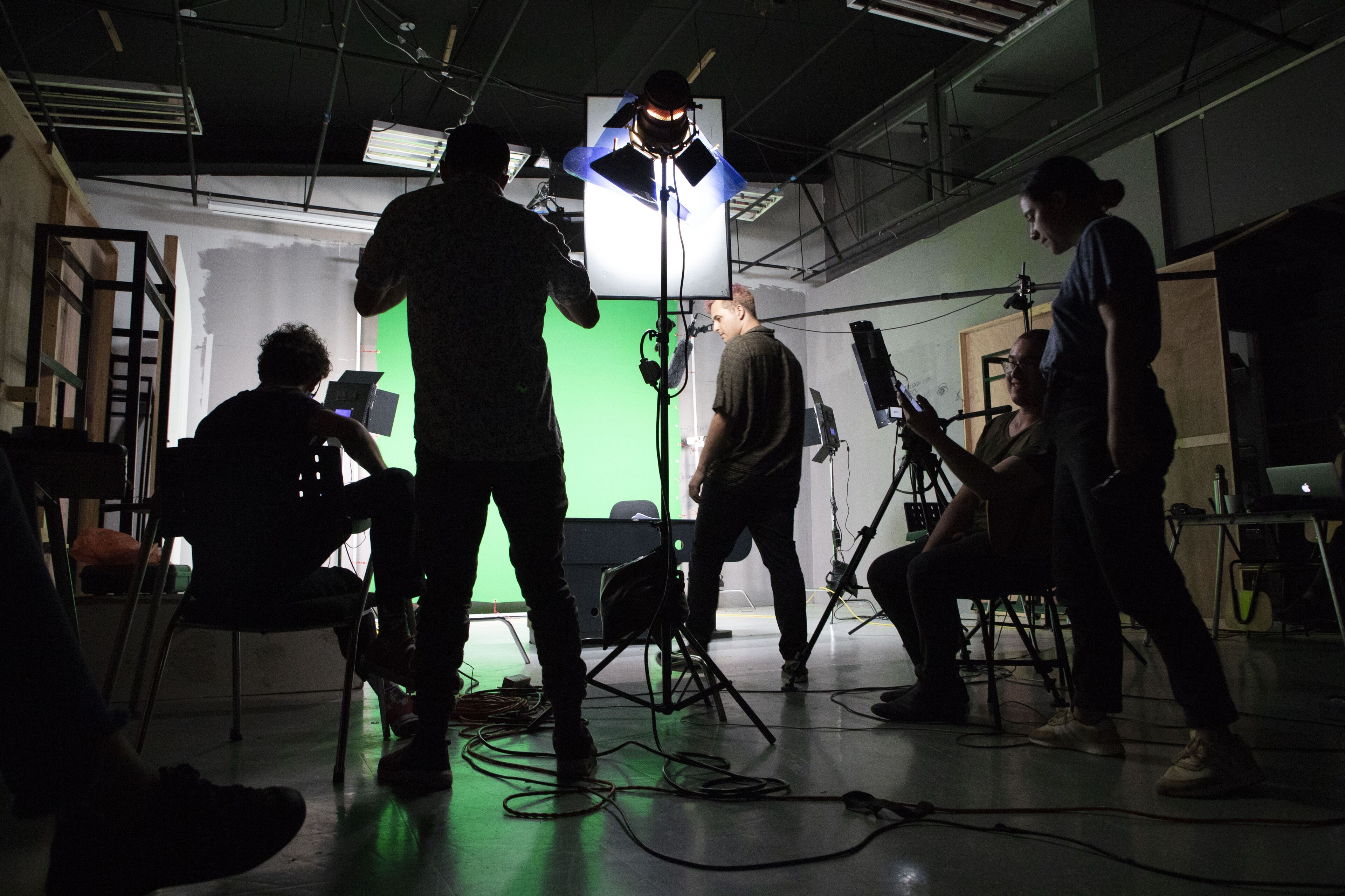 Un equipo de filmación opera con varios equipos en un estudio, silueteados contra luces brillantes y una pantalla verde.