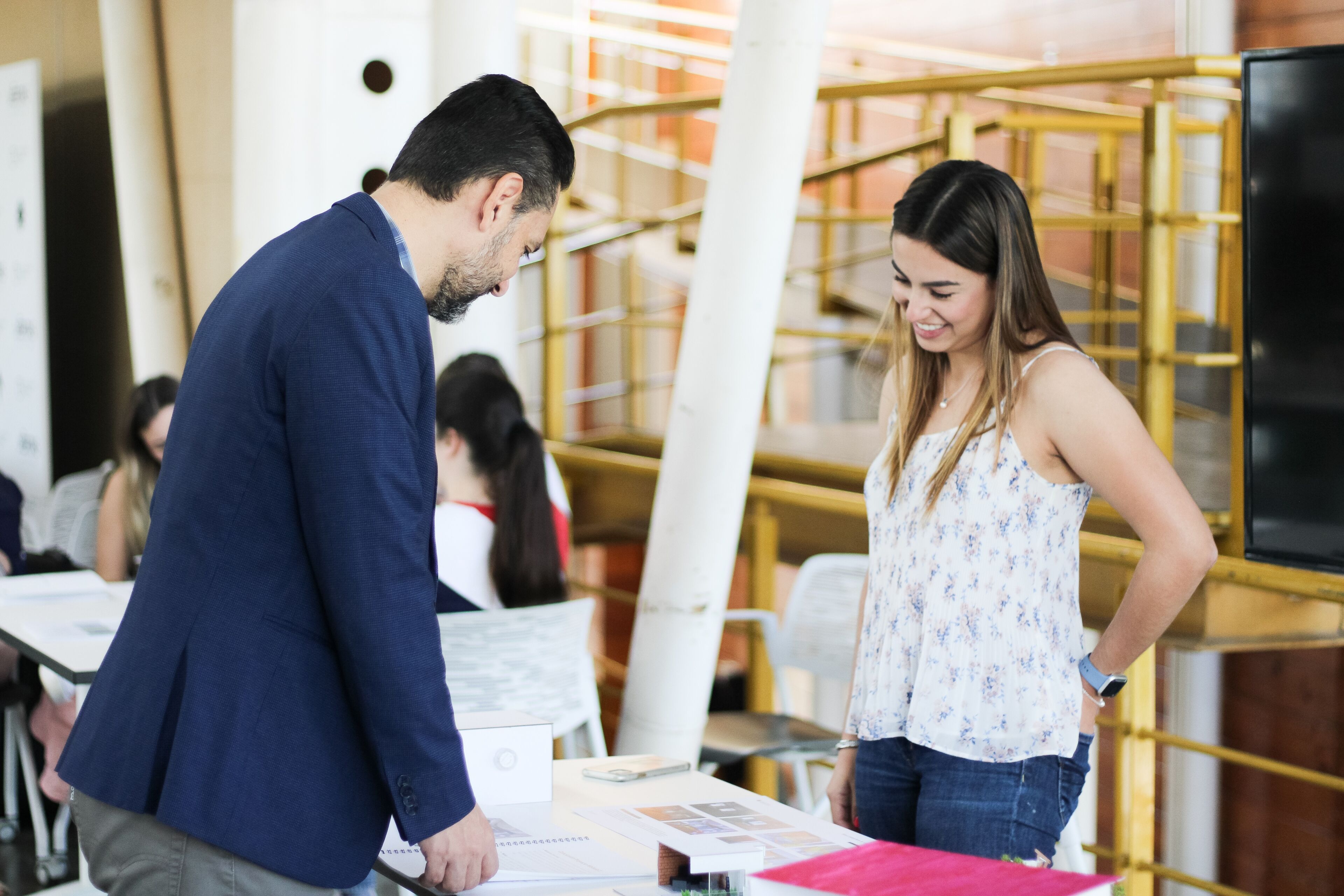 A man in a dark suit examines documents at a conference booth as a smiling woman attendant assists.

