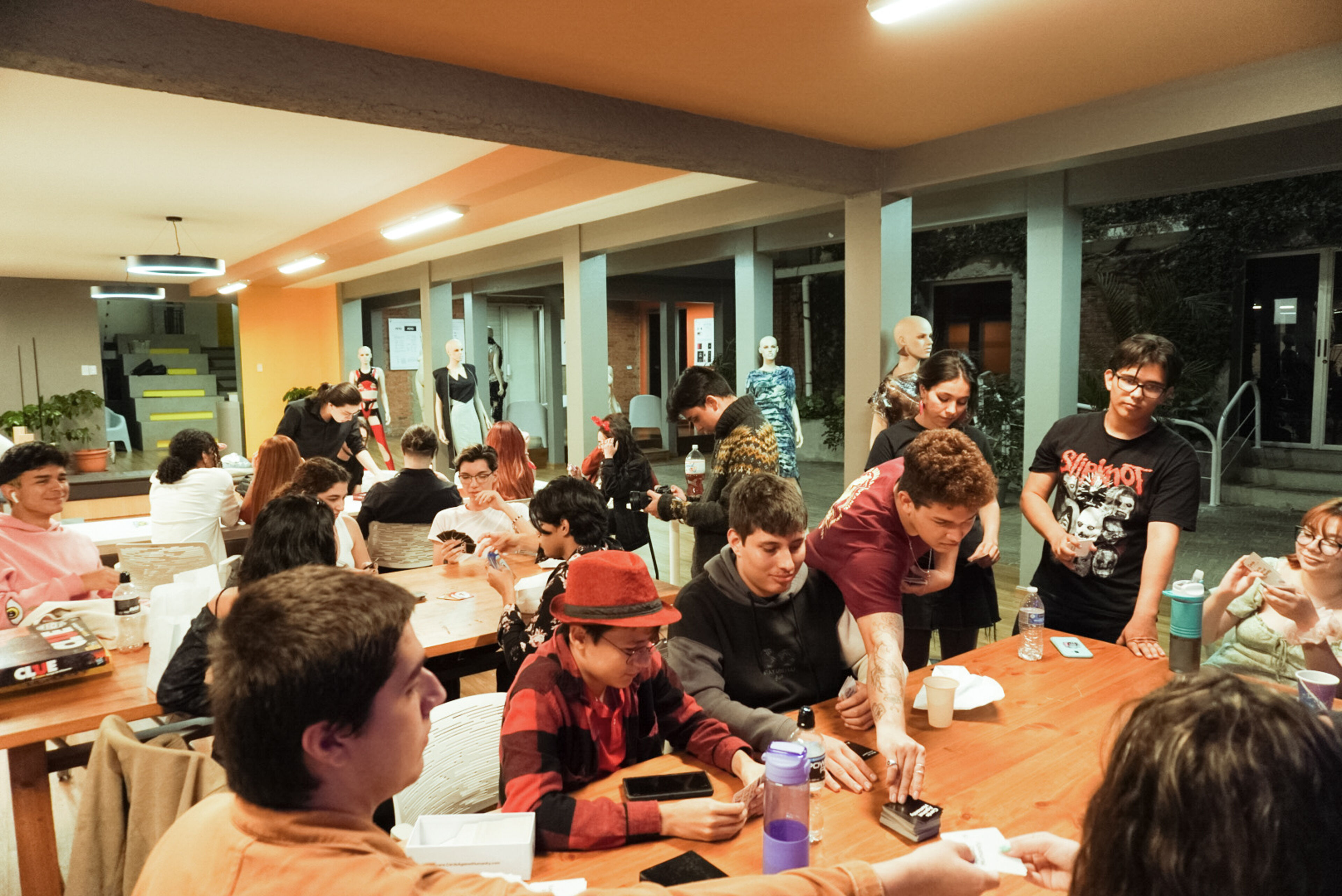 A group of students engage in conversation and card games at a brightly lit campus lounge in the evening.