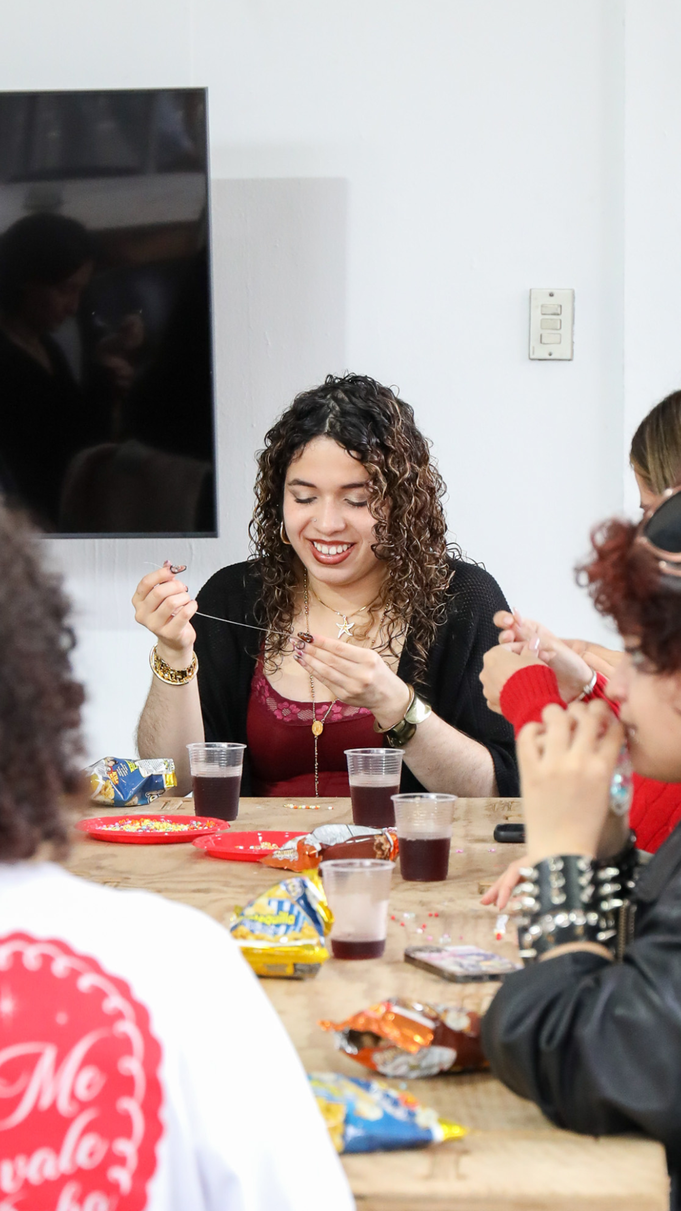 A group of people are gathered around a wooden table, enjoying snacks and drinks. The woman in the center is smiling and holding a small object, possibly a piece of food or jewelry. The atmosphere appears relaxed and social.