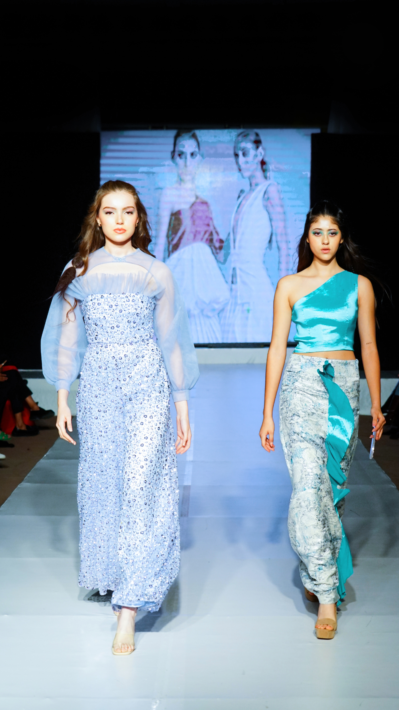 Two models are walking on a runway. The model on the left is wearing a long blue dress, and the model on the right is wearing a blue top and a patterned skirt.