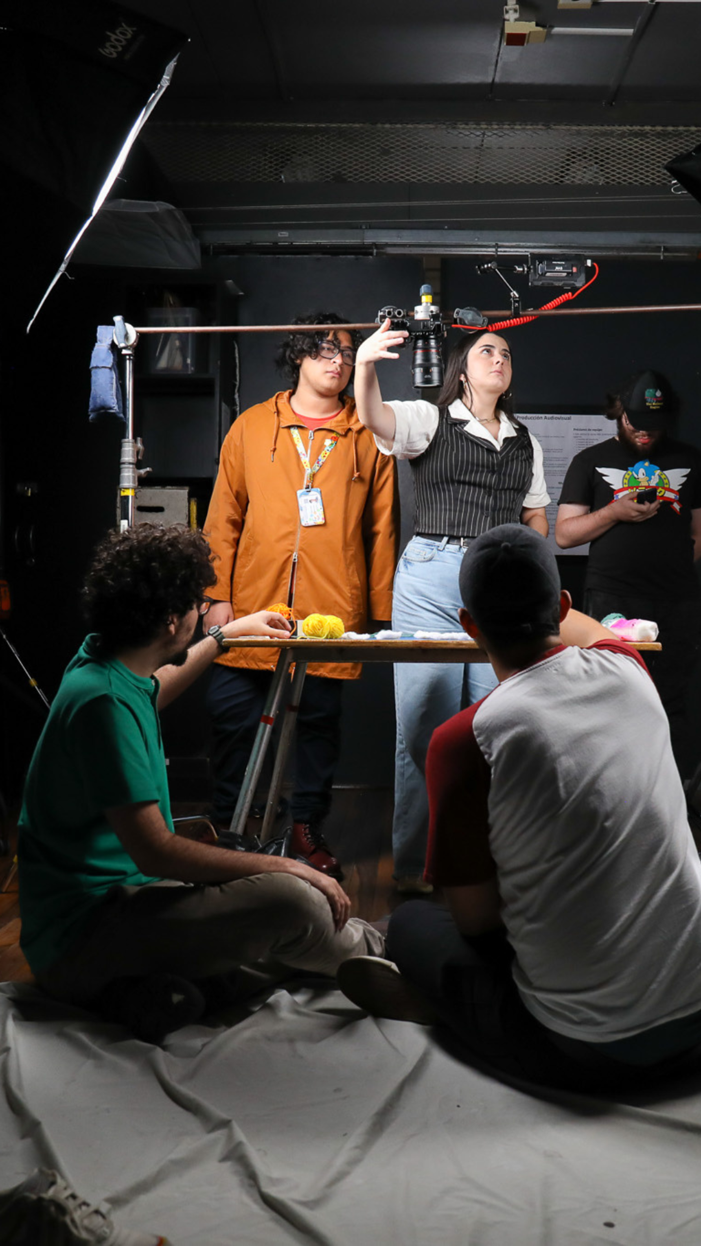 A group of five people are gathered around a table in a studio setting. They appear to be working on a project, possibly involving food items on the table, under studio lighting. The atmosphere seems collaborative and focused.