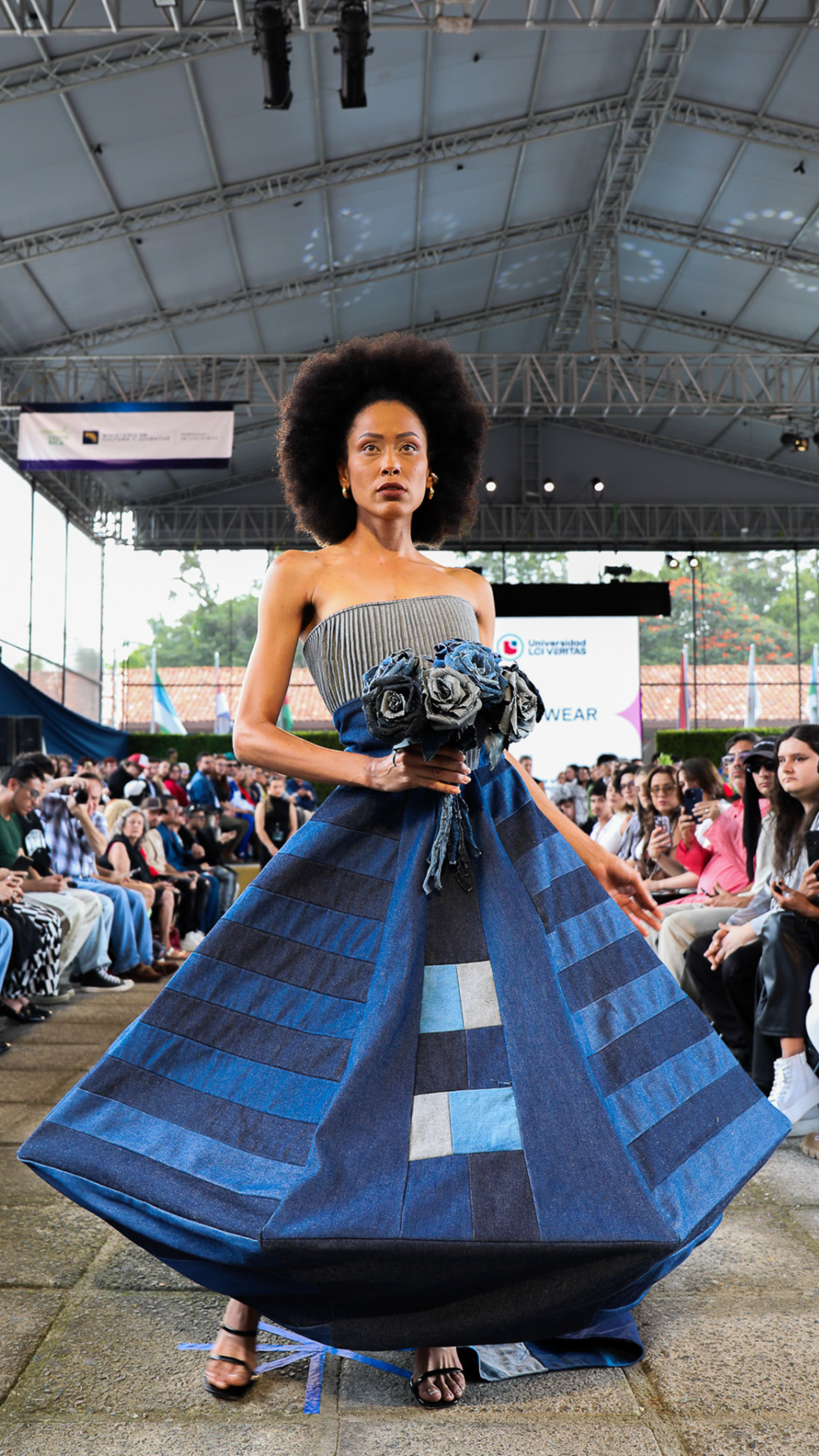 A model is walking down a runway wearing a strapless dress with a gray top and a blue denim skirt. She is holding a bouquet of blue flowers and wearing black heels. The audience is watching her. 