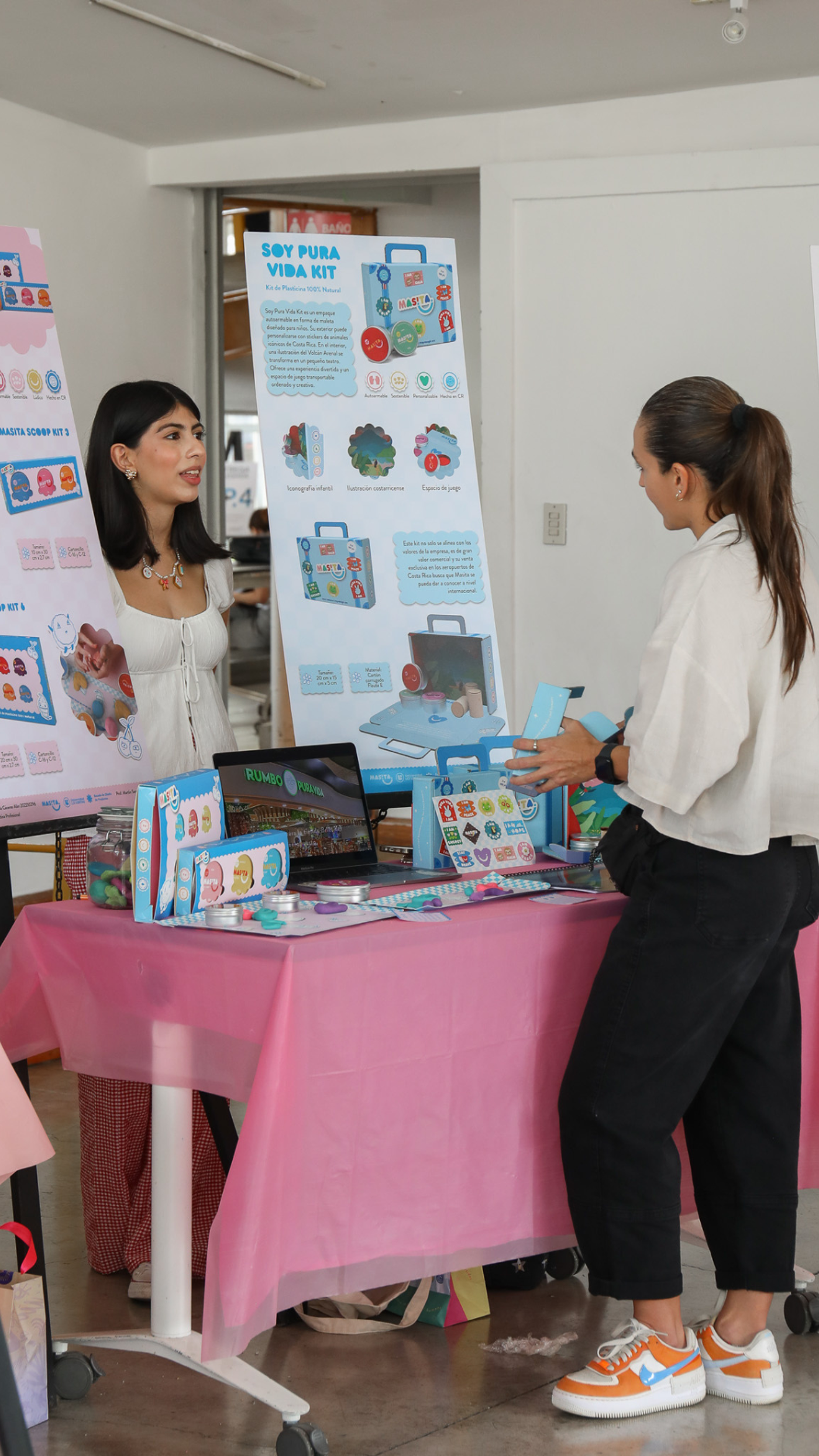 Two women stand at a table covered with a pink tablecloth, displaying a product called "Soy Pusa Vida Kit." One woman is facing the viewer, while the other is facing her, seemingly in conversation. A poster behind them showcases the product details.