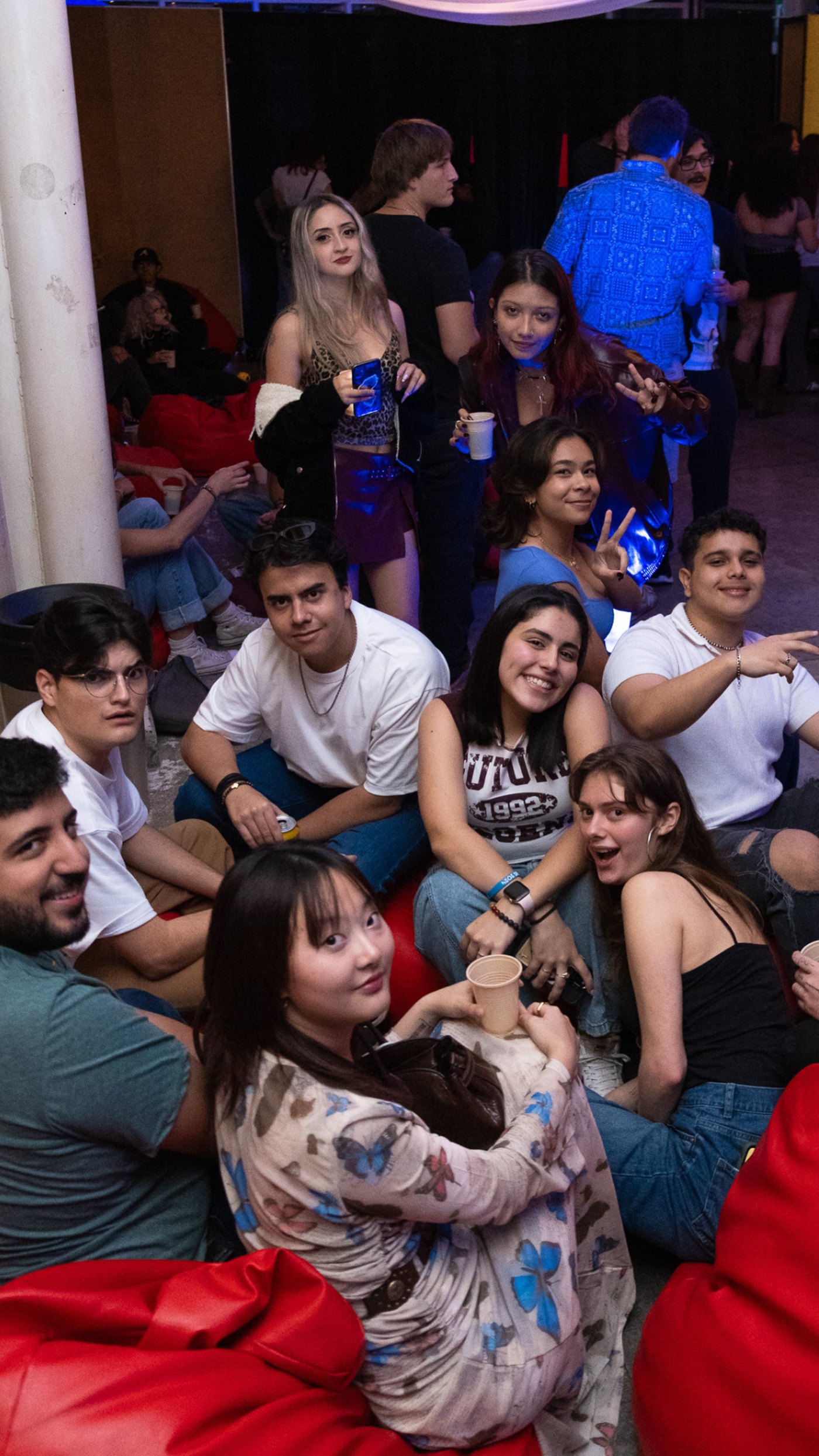 A group of young adults are gathered in a casual setting, possibly a lounge or event space. They are sitting and standing, smiling, and appear to be enjoying each other's company in a relaxed atmosphere.