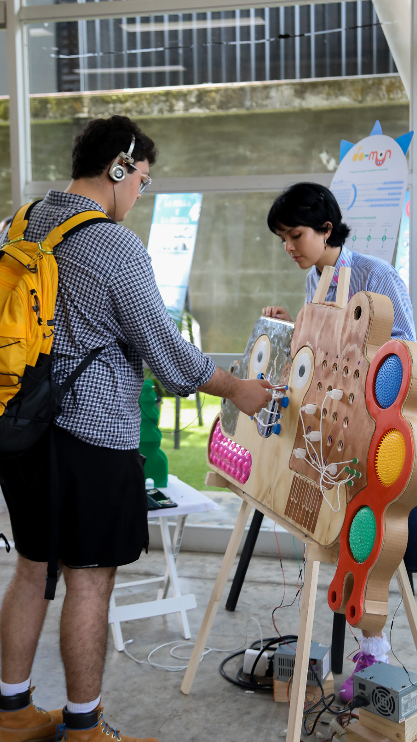Two people are interacting with a large, colorful interactive display. One person is wearing headphones and a backpack, while the other is focused on the display. The display features various buttons, lights, and textures, suggesting it's designed for sensory exploration.