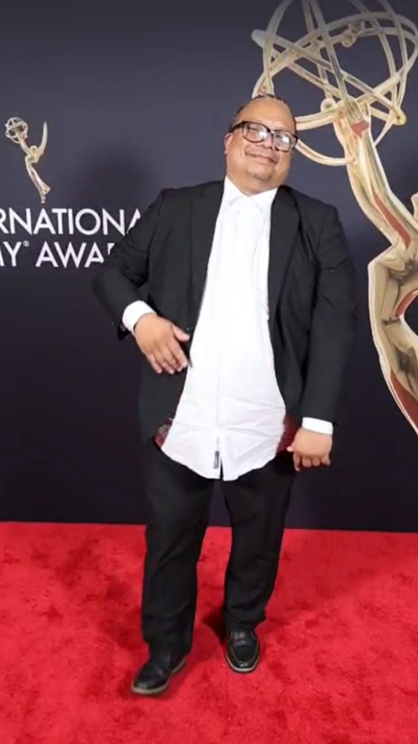 The image shows a man in a suit posing on a red carpet. He is smiling and appears to be at an awards ceremony. The background includes an award logo.