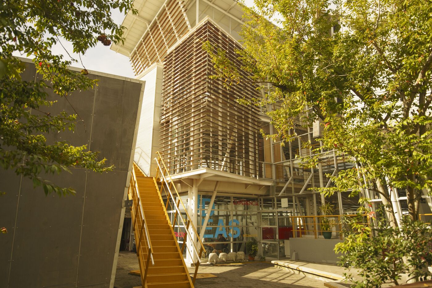 A contemporary eco-friendly building with a vertical garden, solar panels, and a striking yellow staircase, nestled among lush greenery in an urban setting.