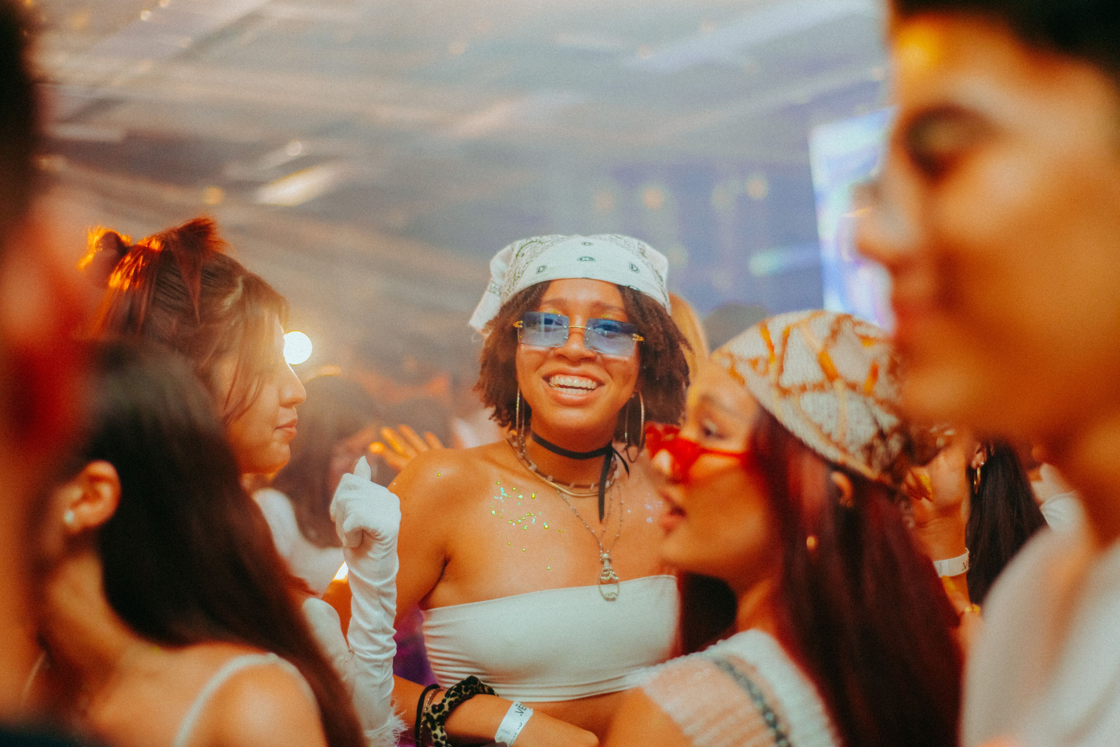 Una joven alegre con top blanco y bandana sonríe brillantemente, rodeada de amigos en una fiesta vibrante.