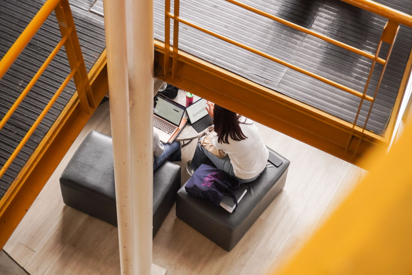 A student focuses on her laptop while seated on a dark cushioned bench in a well-lit library with striking yellow stairs.
