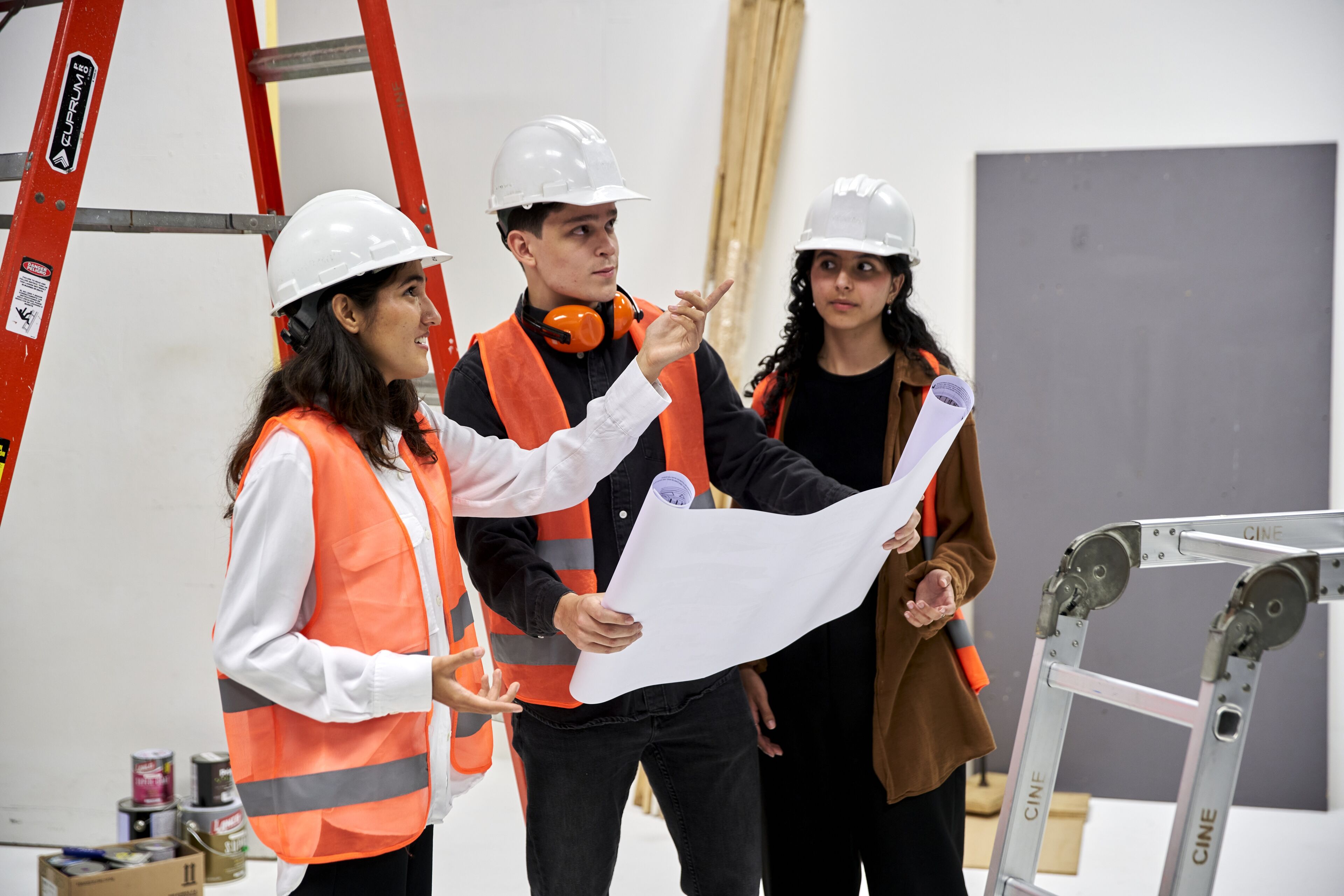 Three professionals in safety gear with hard hats and high-visibility vests discuss over a blueprint in a construction site.