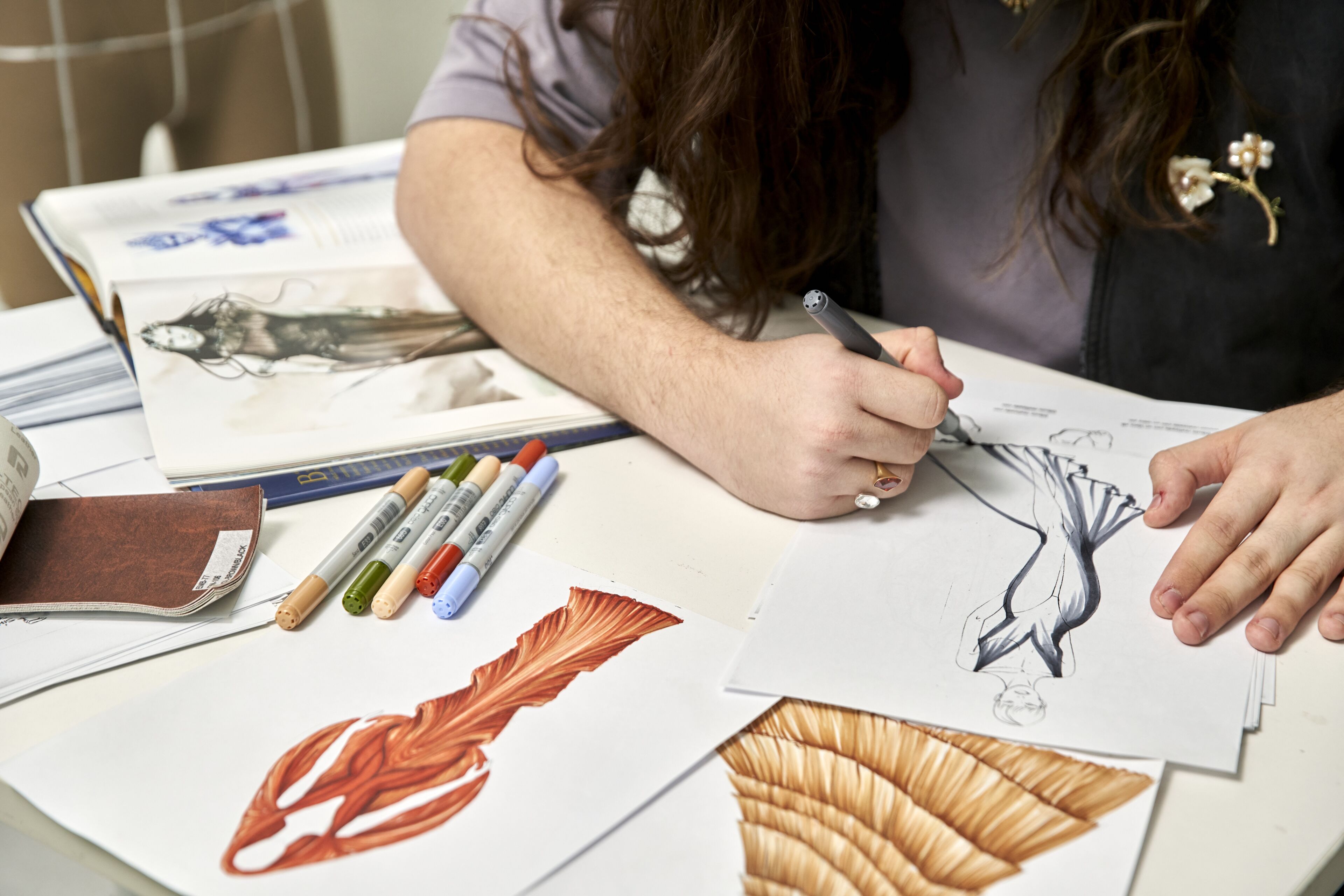 An individual sketches a dress design on paper, surrounded by markers and various drawing materials.