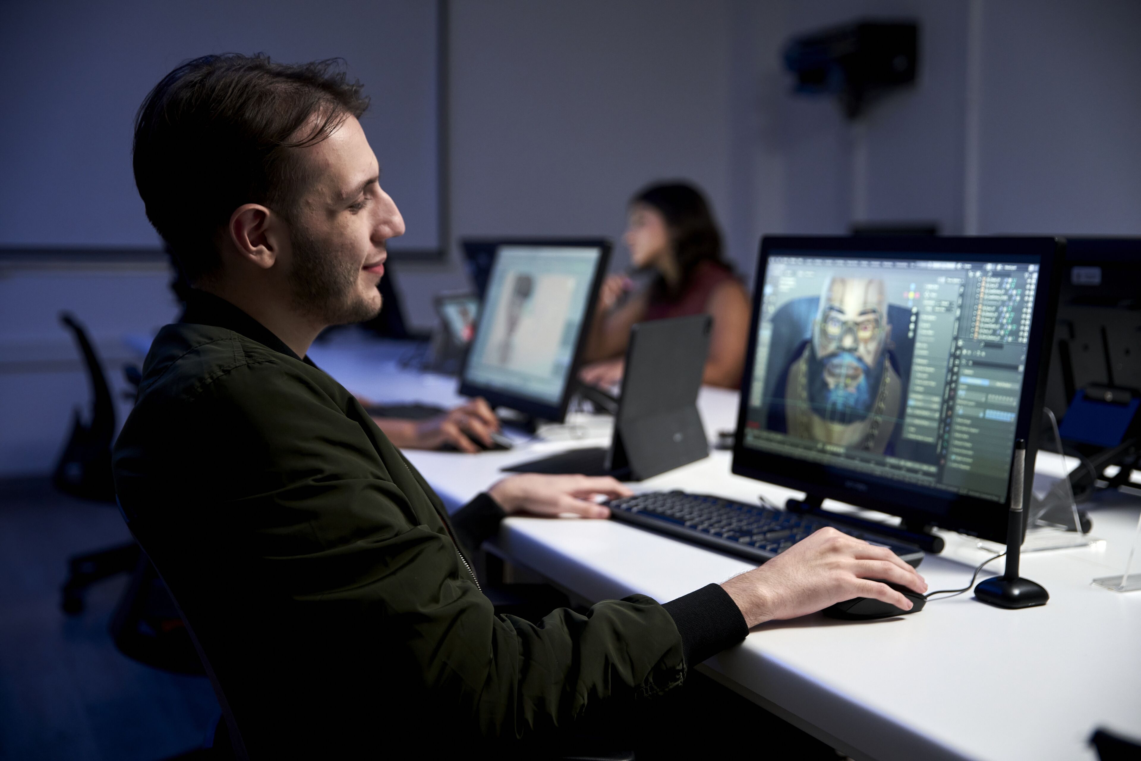A focused male 3D artist works on a character model in a dimly lit digital animation studio.