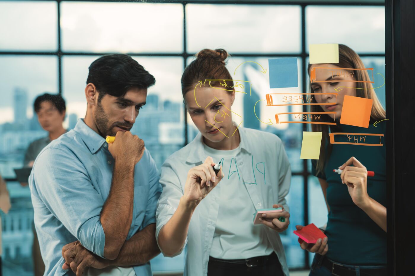 Three people are collaborating in an office, writing ideas on glass with markers and sticky notes, planning a project.