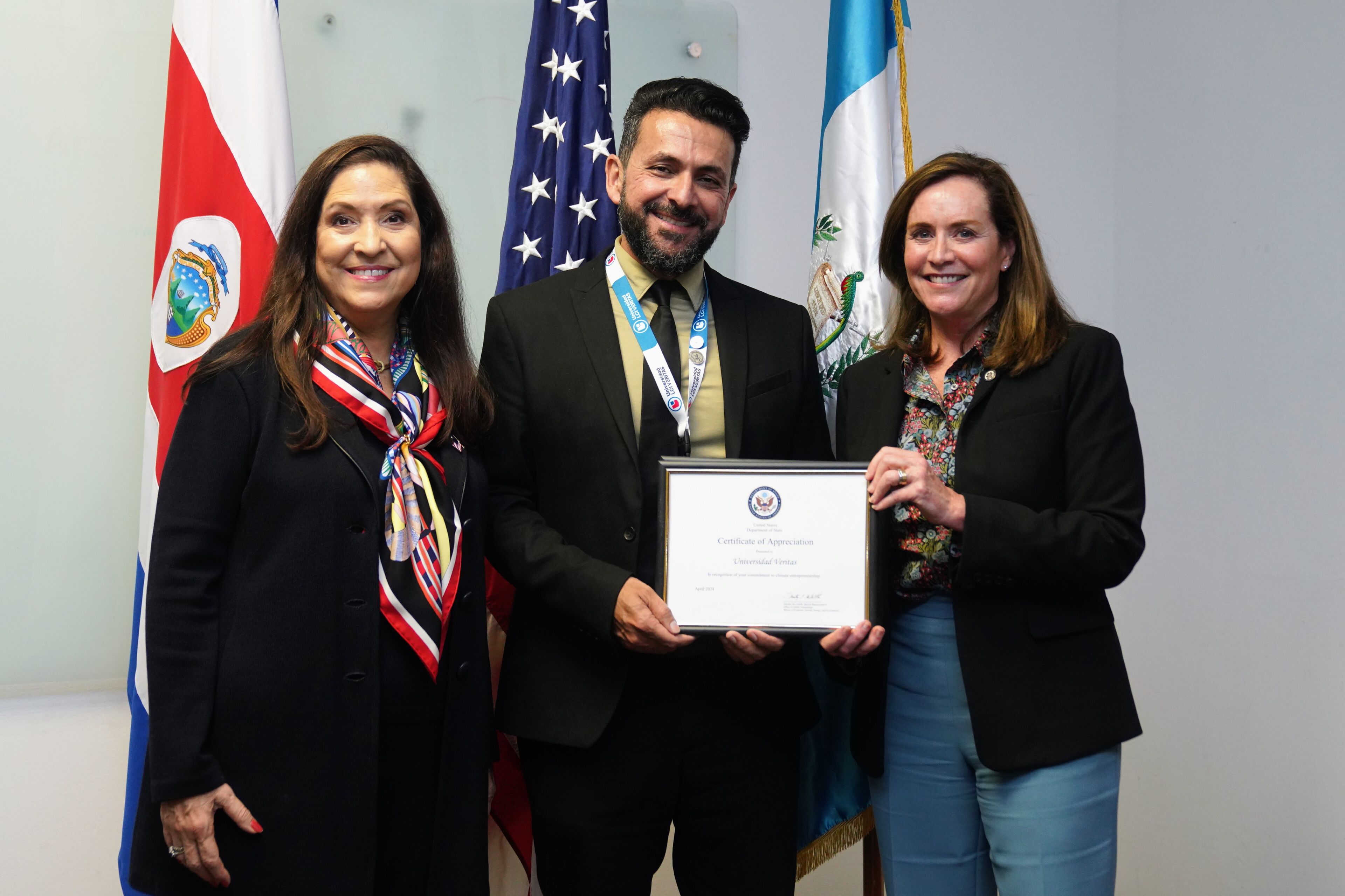 Esta imagen muestra una ceremonia de reconocimiento internacional. Tres personas, incluyendo dos mujeres y un hombre, están juntos, sonriendo. El hombre sostiene un Certificado de Apreciación enmarcado. Detrás de ellos están las banderas de Costa Rica, Estados Unidos y Guatemala, lo que indica el contexto internacional.