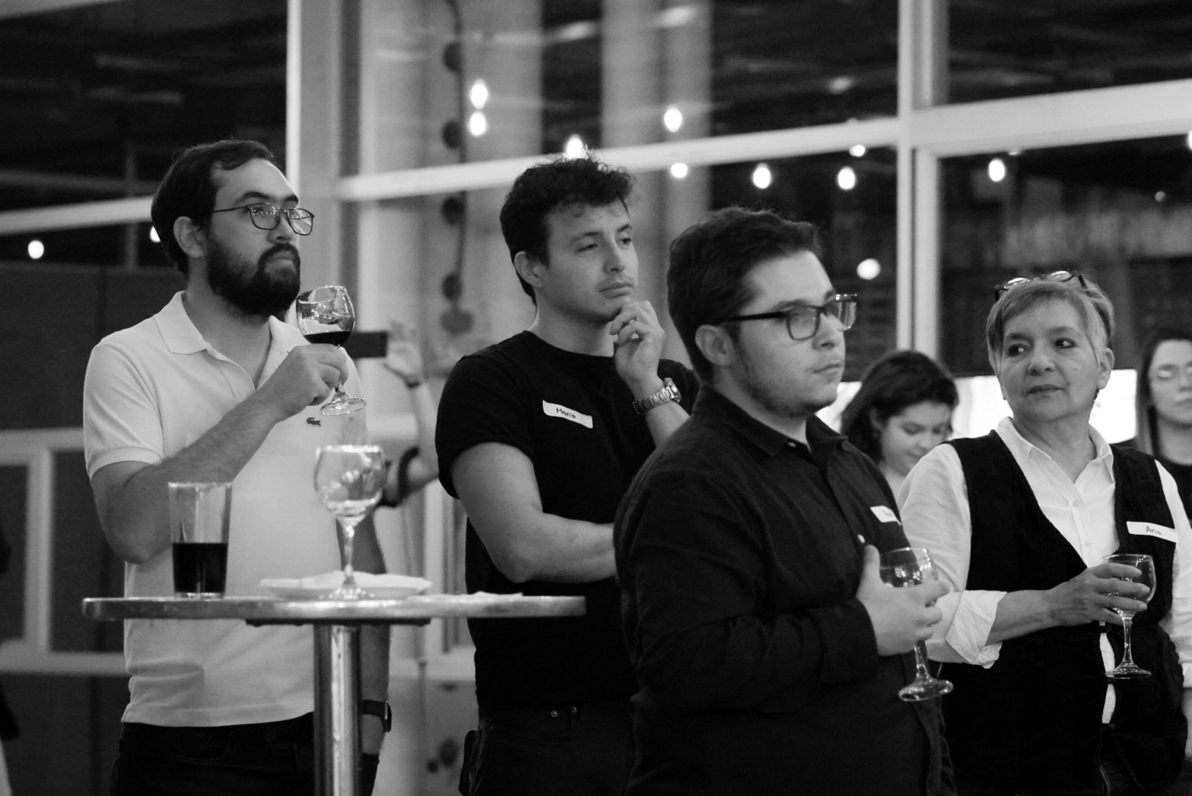 Un grupo de individuos atentos con copas de vino, escuchando con interés en una reunión social.