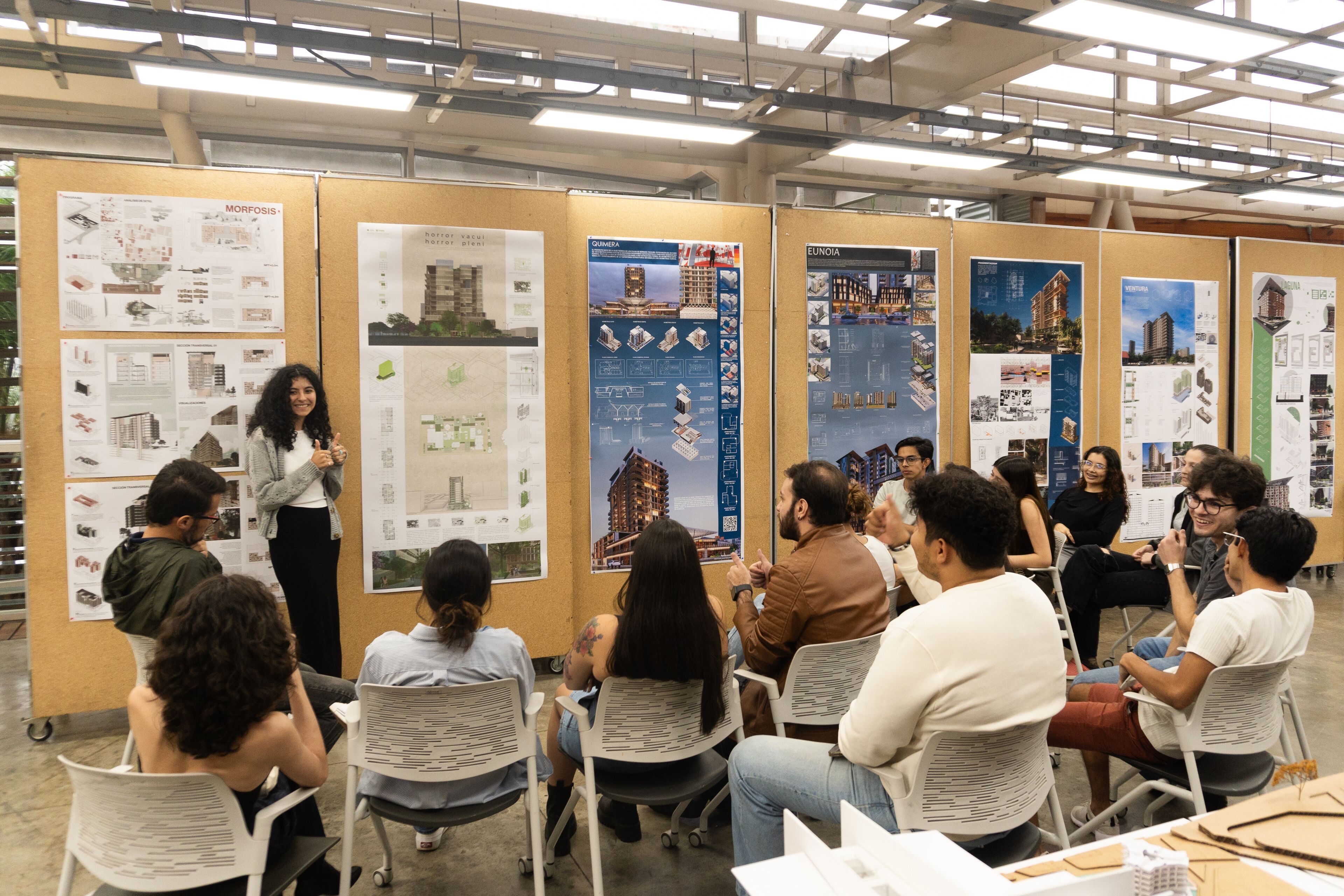 A young woman presents architectural projects to an attentive audience in a well-lit workshop space.