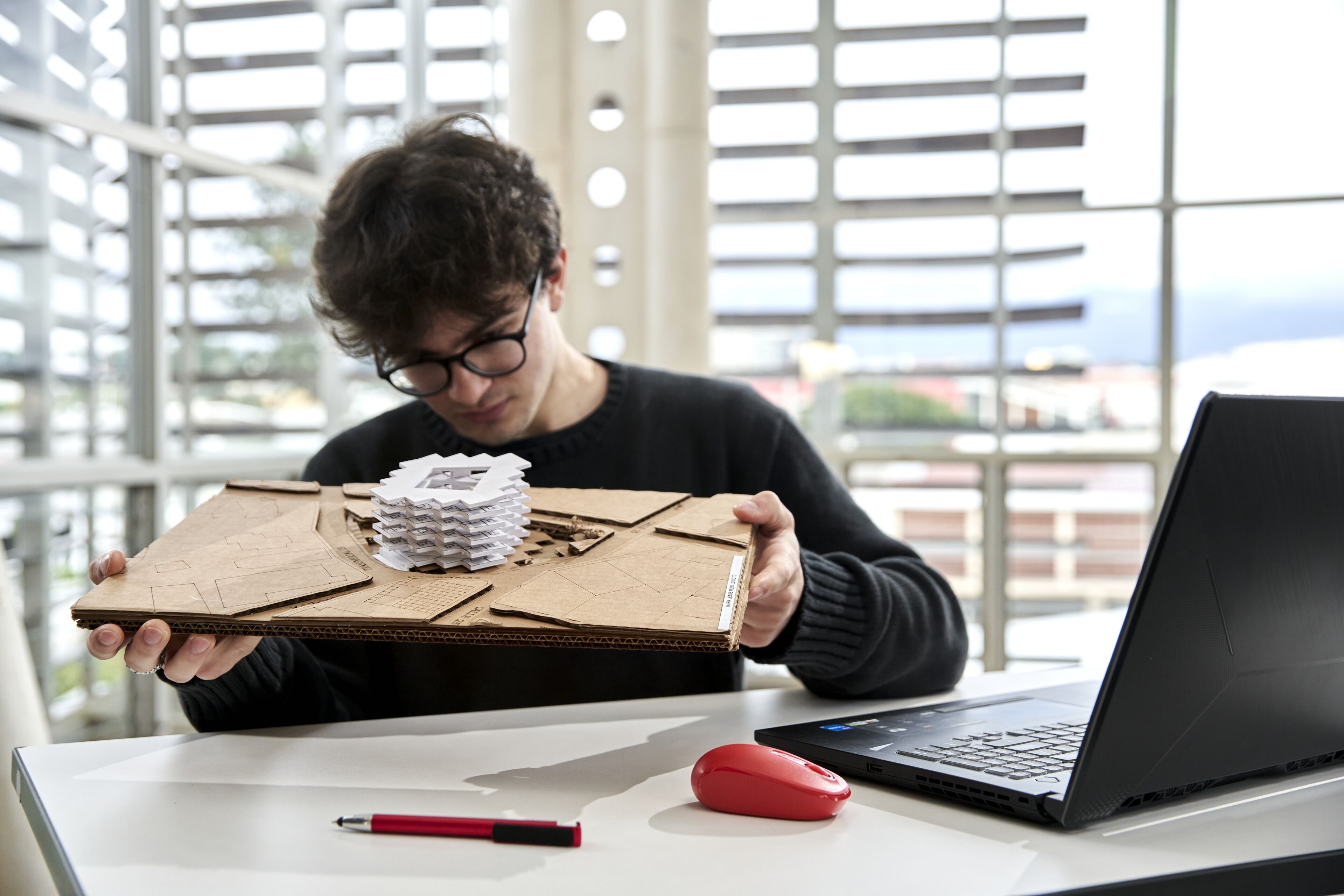 Un jeune architecte examine attentivement un modèle architectural blanc sur un socle en carton, avec un ordinateur portable et une souris rouge sur la table.