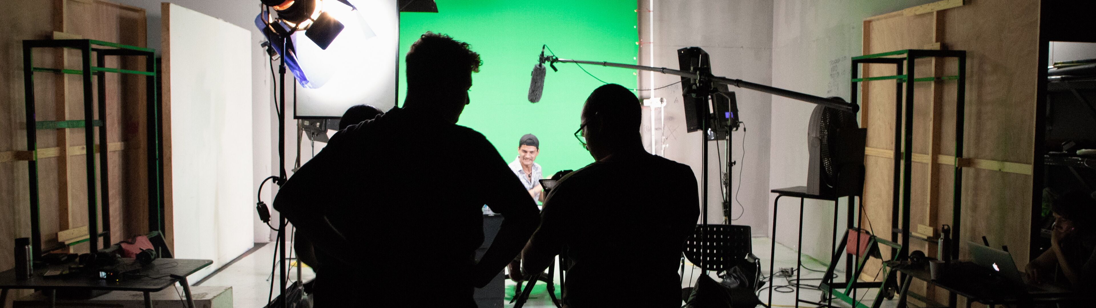 Siluetas de miembros del equipo trabajando en un set de filmación con equipos de iluminación y una pantalla verde.