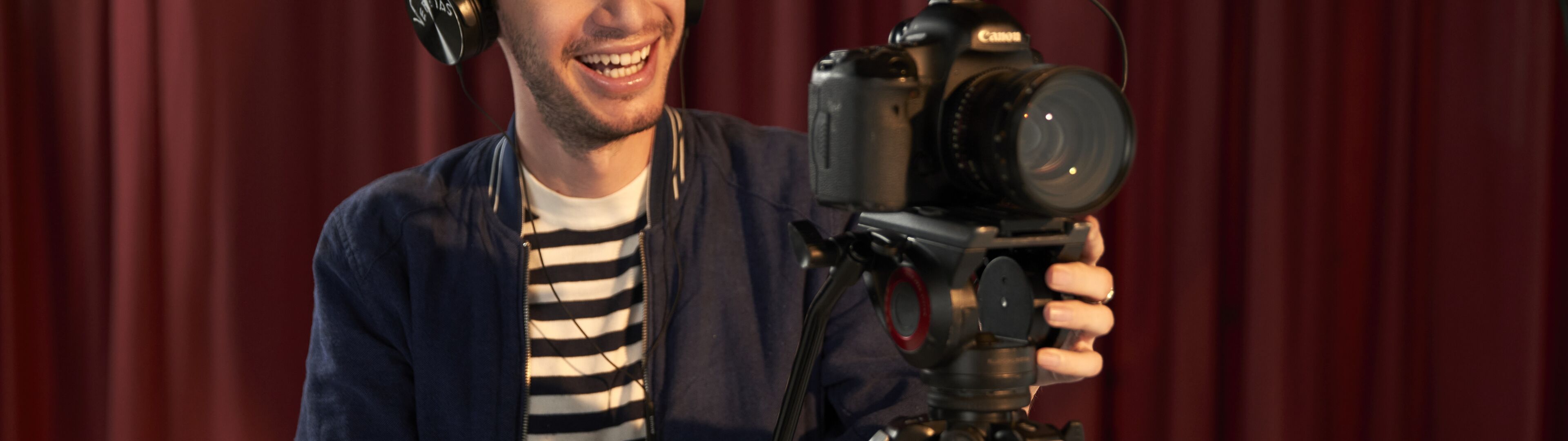 Un hombre sonriente maneja una cámara de vídeo profesional en un trípode, con auriculares, frente a un telón rojo.