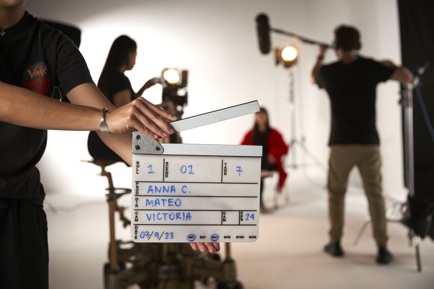 A clapperboard is held in focus on a film set, with crew and lighting equipment in the background.