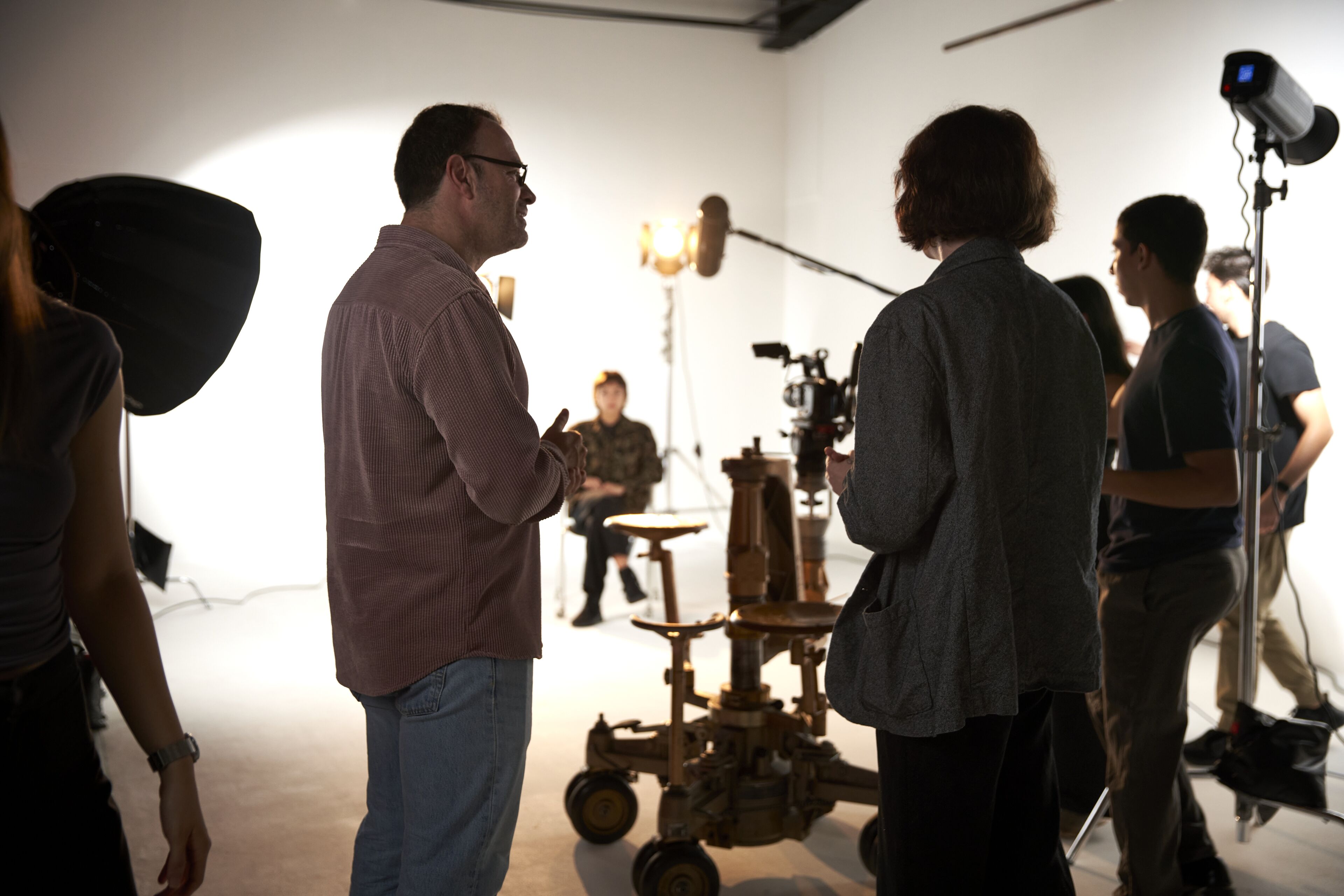 Director conversando con un miembro del equipo en un set de filmación, con equipo de iluminación y cámara al fondo.
