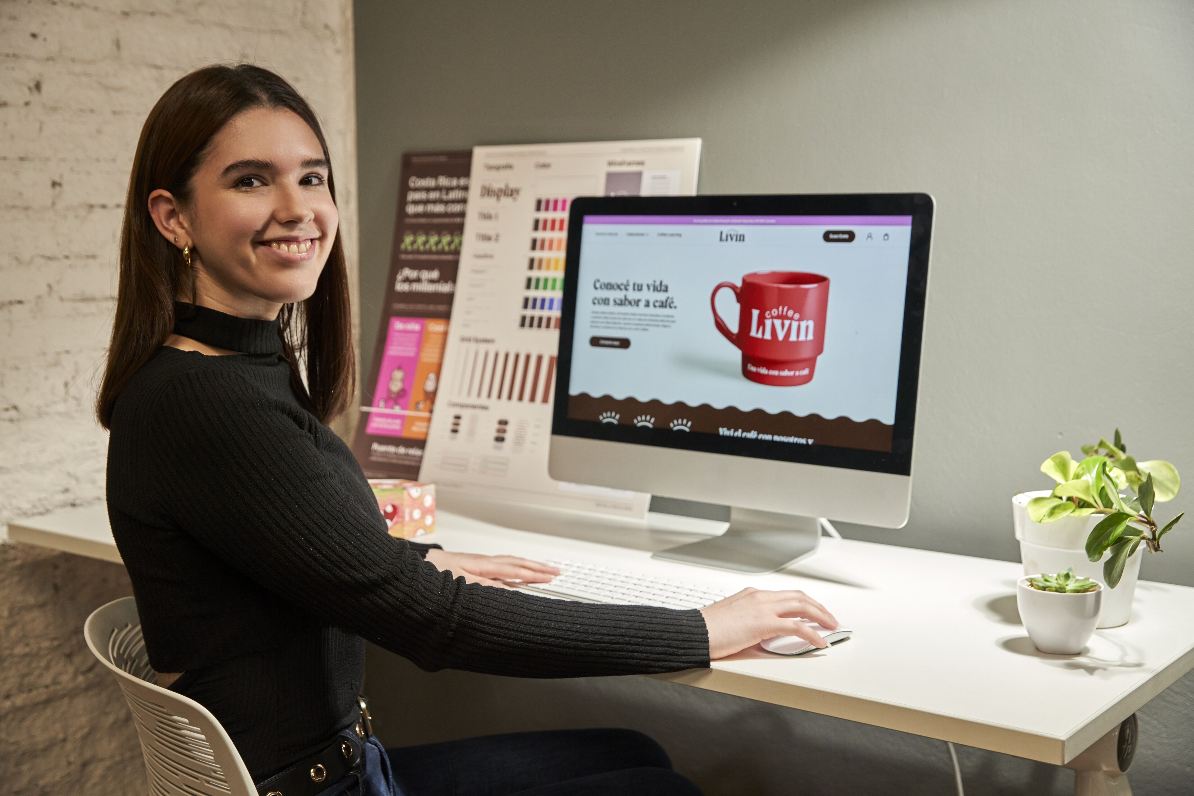Una mujer joven sonriendo a la cámara, sentada en un espacio de trabajo moderno con un ordenador que muestra un anuncio de café.