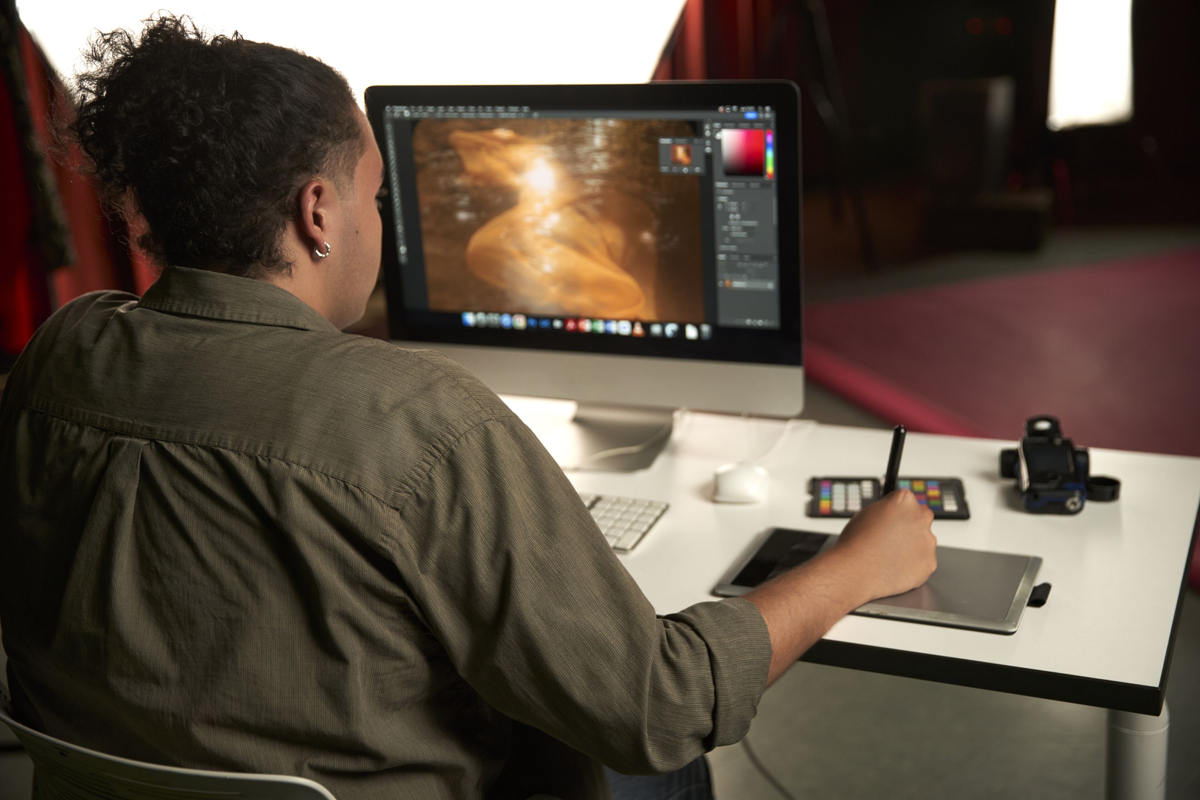 Un diseñador gráfico está concentrado en editar una imagen en un ordenador, con una tableta gráfica y muestras de color en el escritorio.