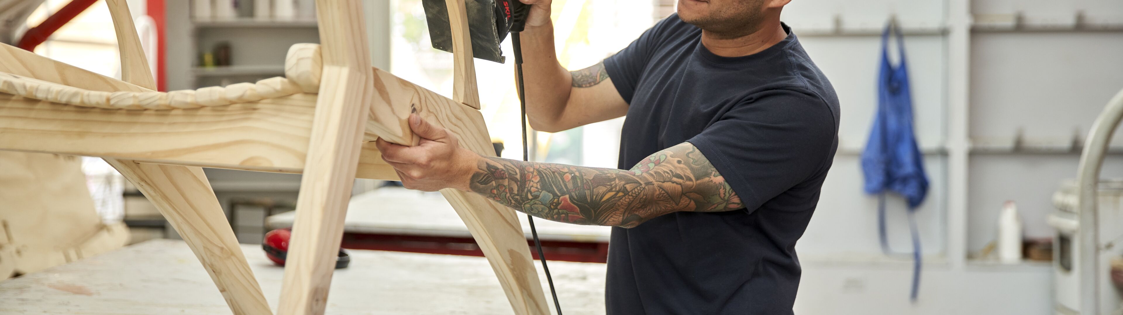 Un artesano tatuado con gafas de seguridad lija una silla en un taller bien iluminado.