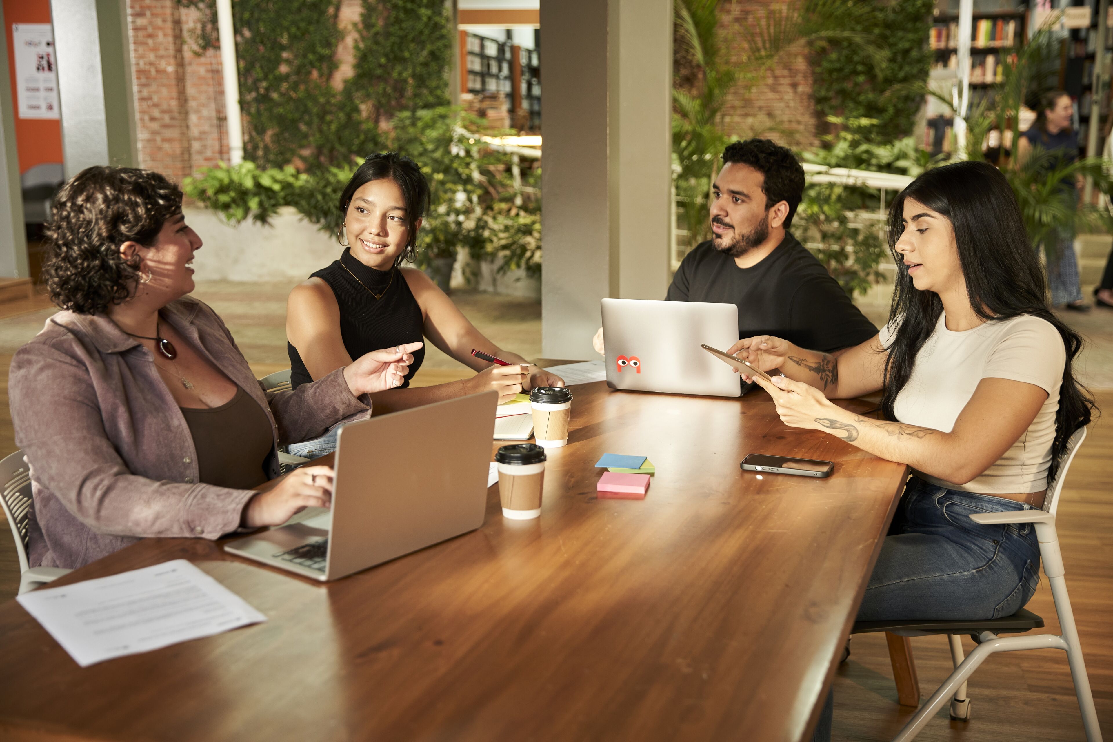 Cuatro profesionales en una reunión colaborativa, con portátiles y blocs de notas, en una mesa de madera en un entorno de co-working moderno.