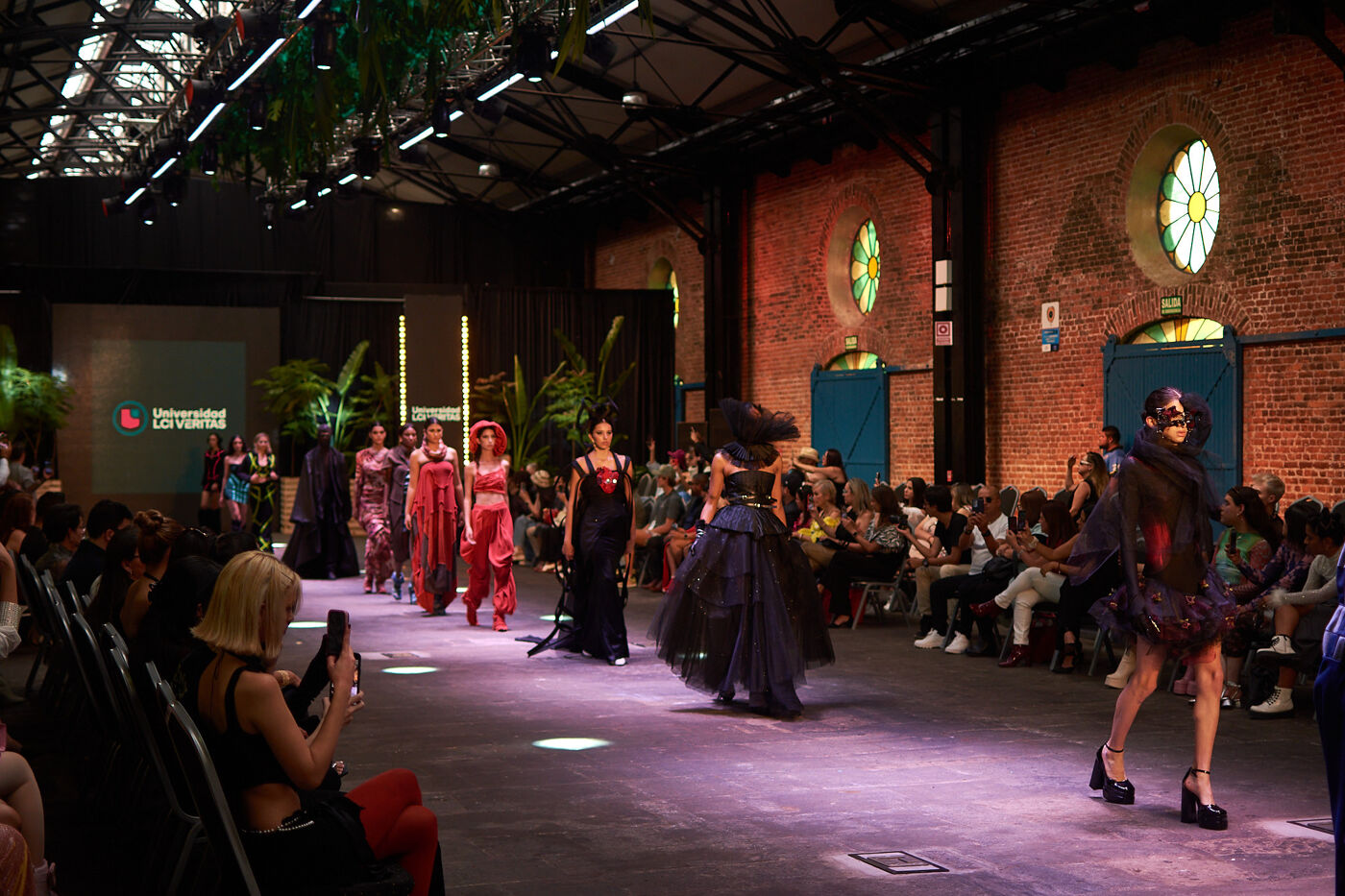 Models parading contemporary designs down a runway at a fashion show hosted by Universidad La Verdad, with an audience capturing the event on their phones.