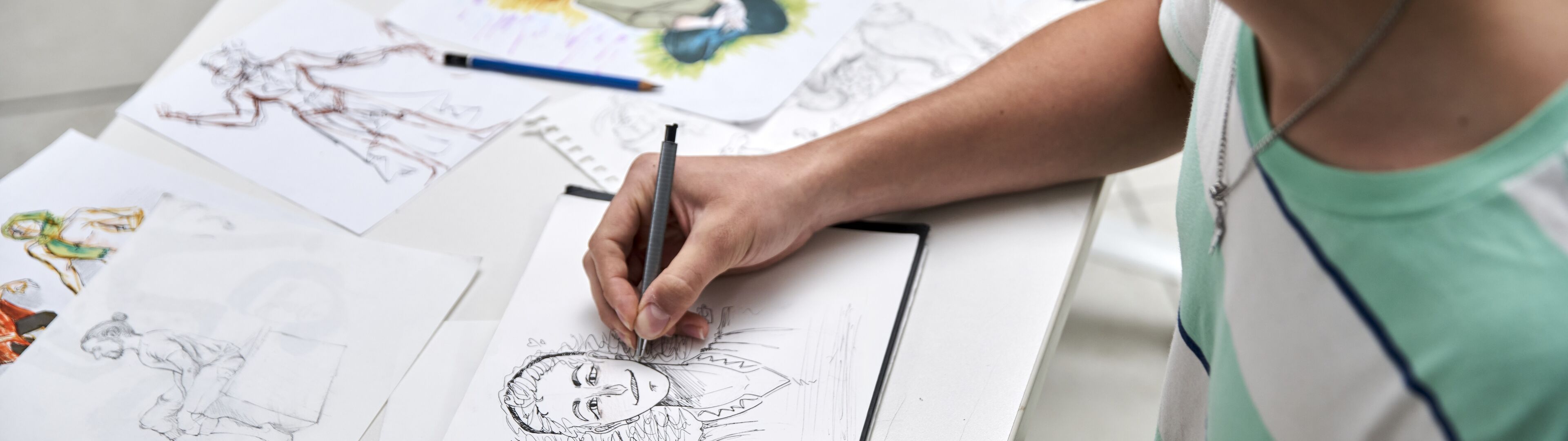 Una persona concentrada dibujando un retrato detallado entre varios bocetos en un escritorio.