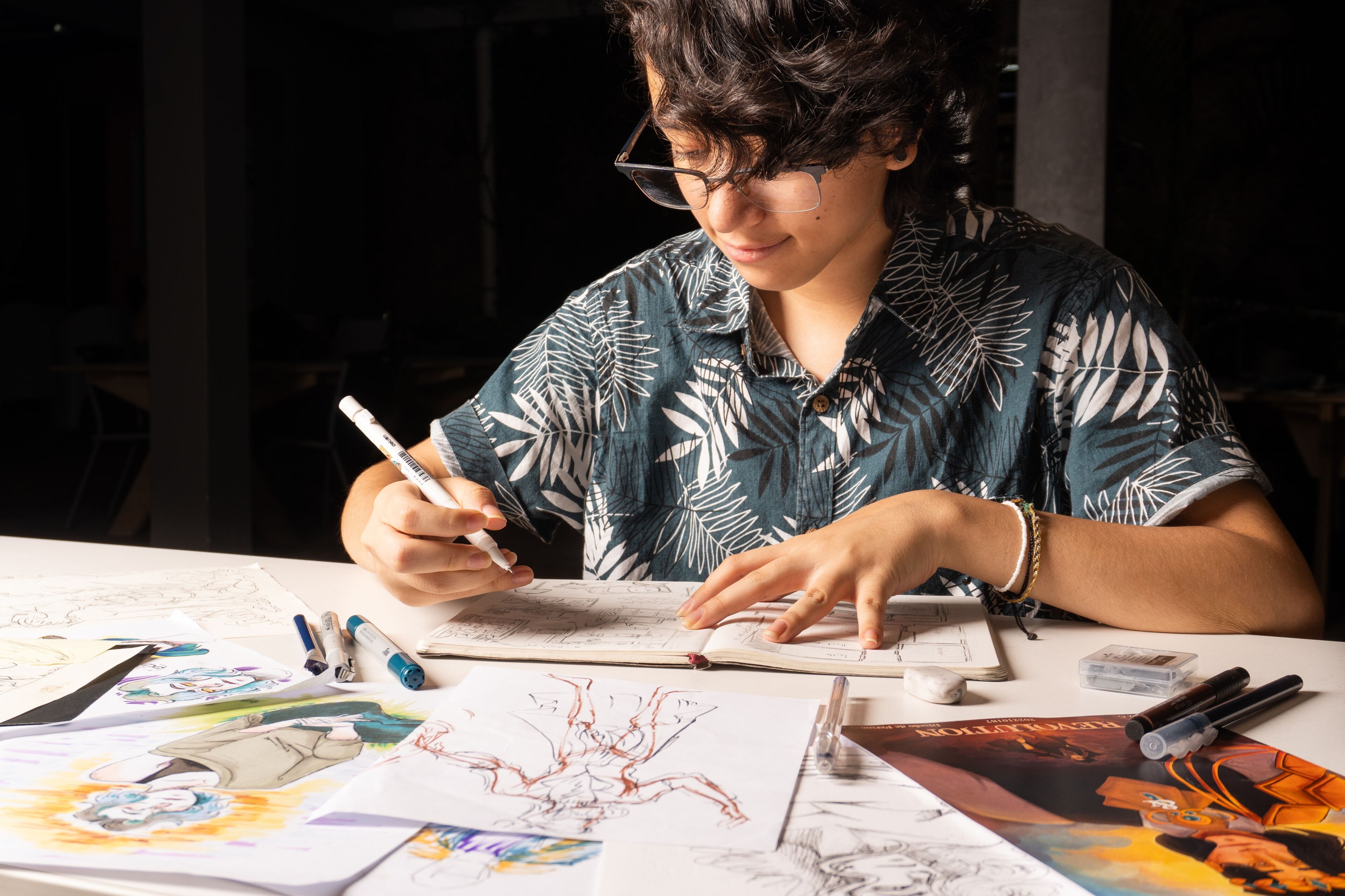 Una persona enfocada boceta ilustraciones de personajes, rodeada de materiales de arte y dibujos.