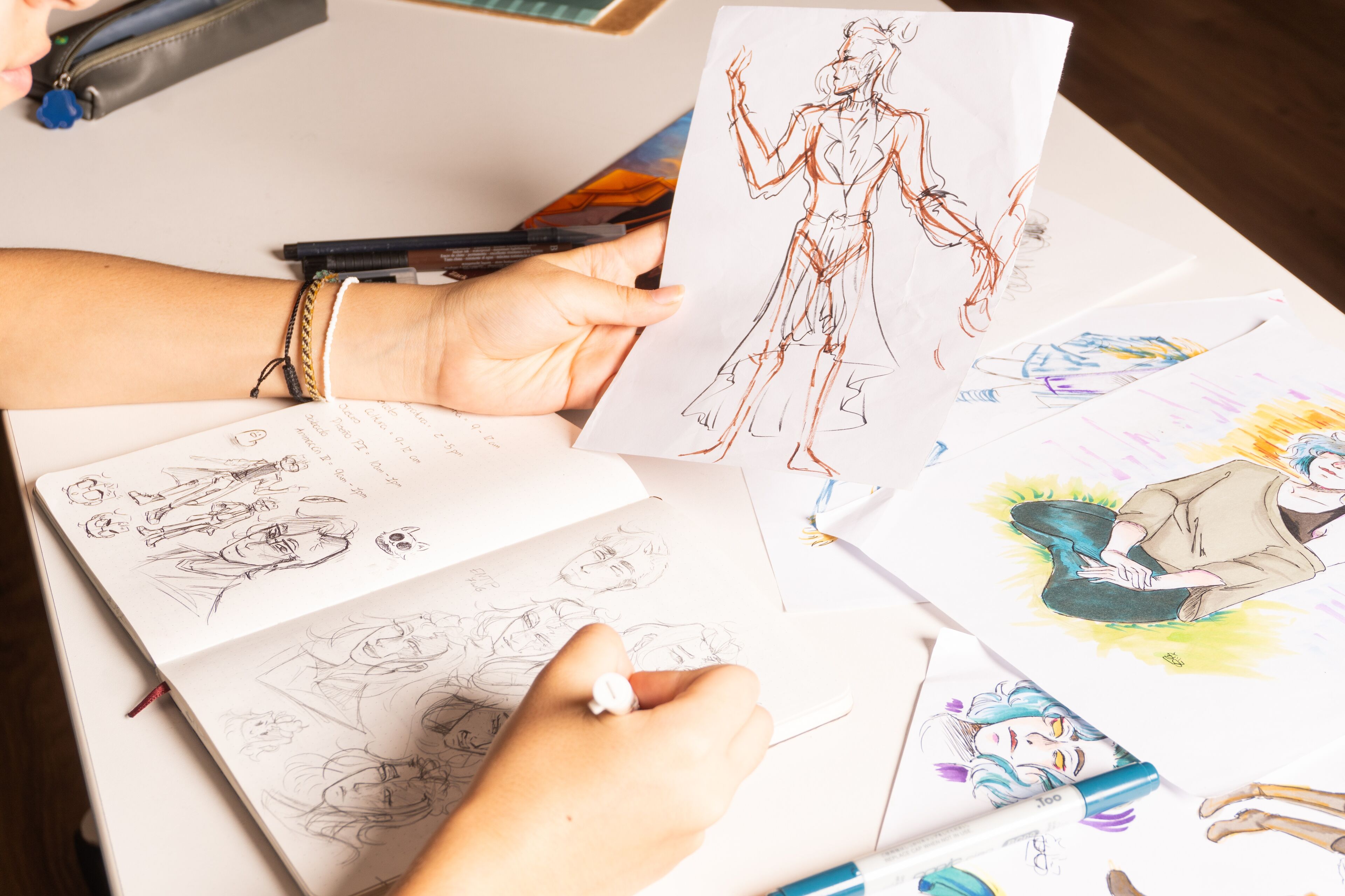 La mano de un artista esboza en un cuaderno rodeado de dibujos de personajes dispersos.