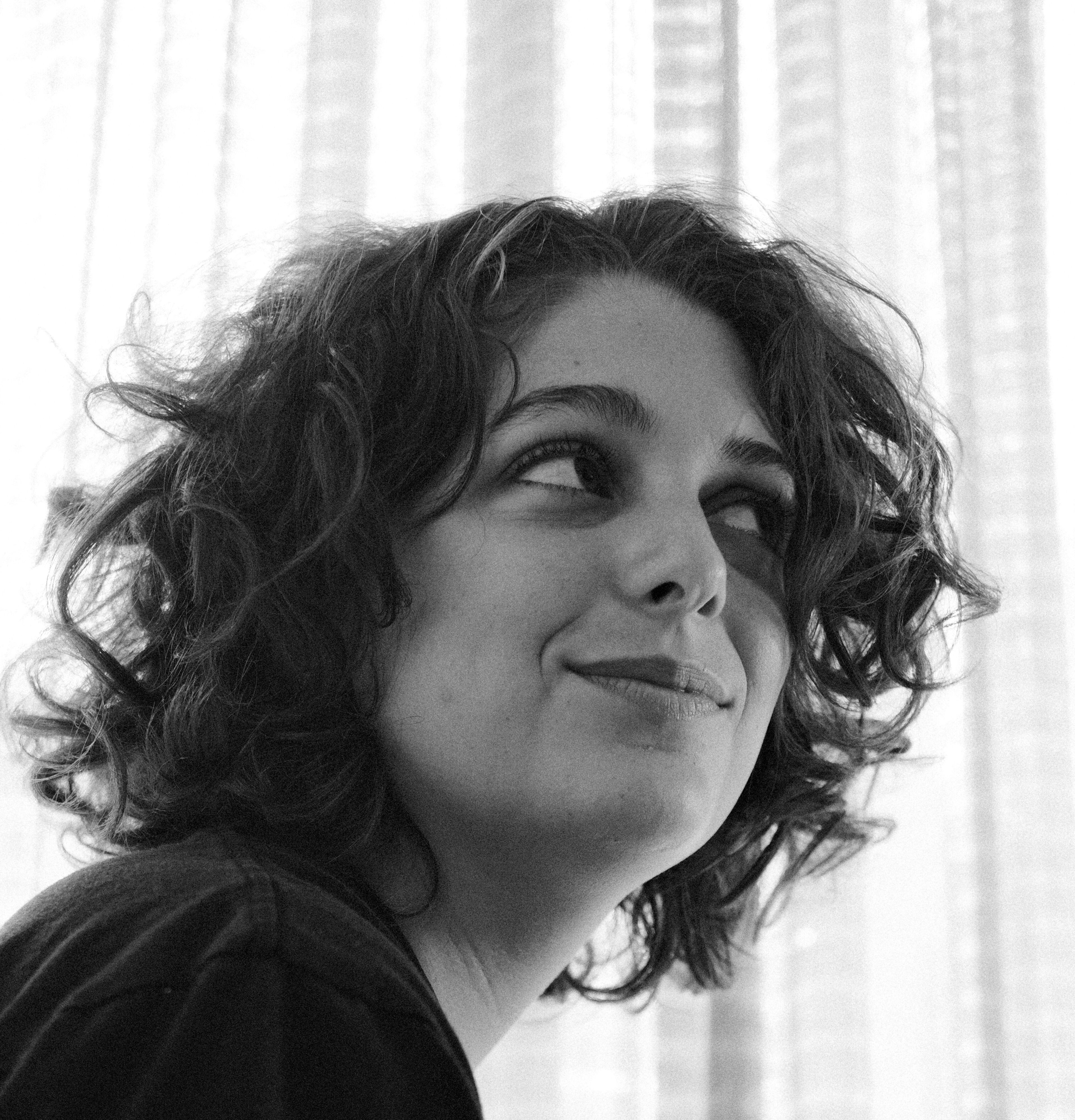 Retrato en blanco y negro de una mujer joven con cabello rizado, sonriendo sutilmente mientras mira hacia el lado, con cortinas translúcidas difuminando la luz de fondo.