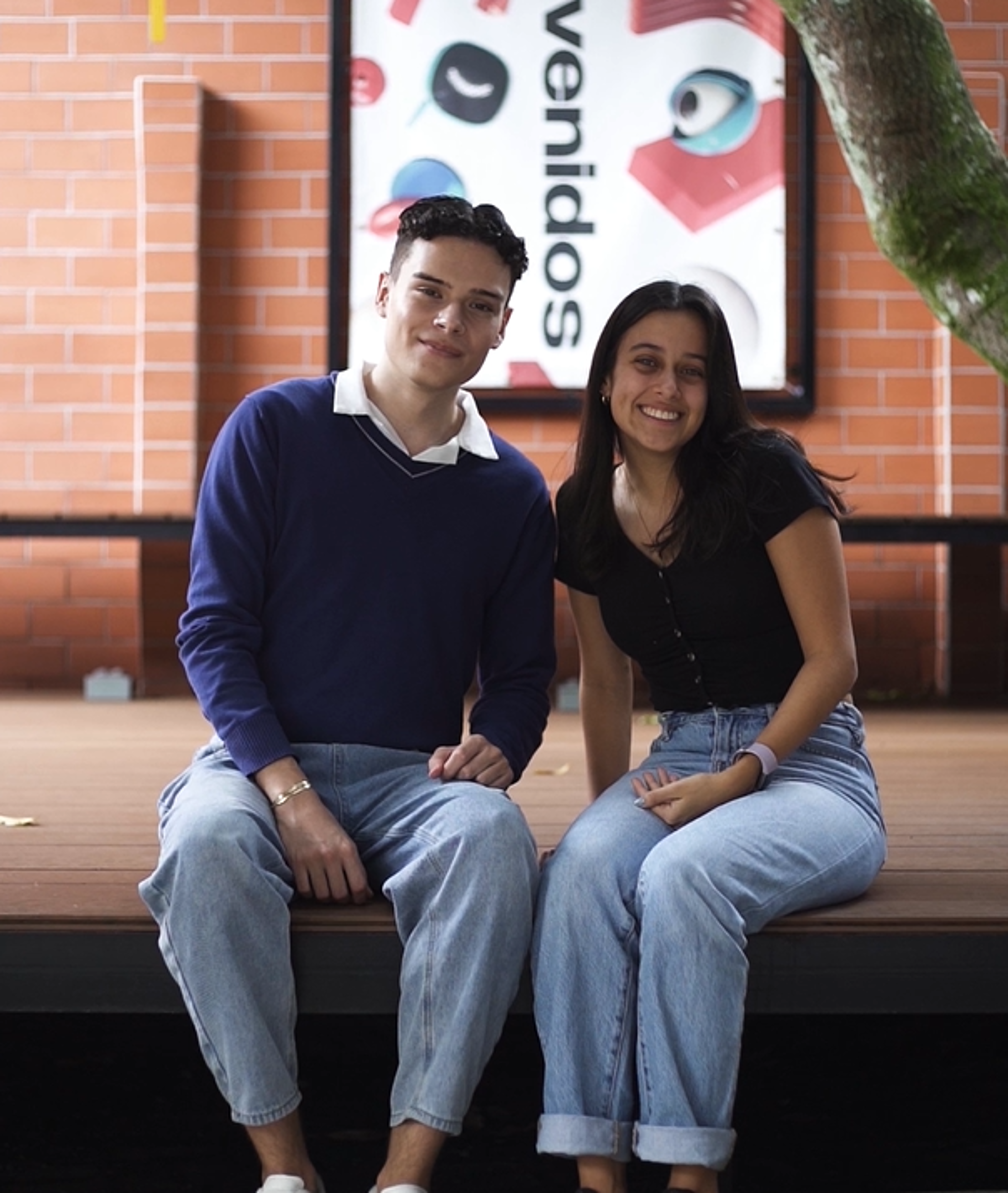 Un joven y una joven sentados sonrientes en una banca, con un cartel de bienvenida en español de fondo.